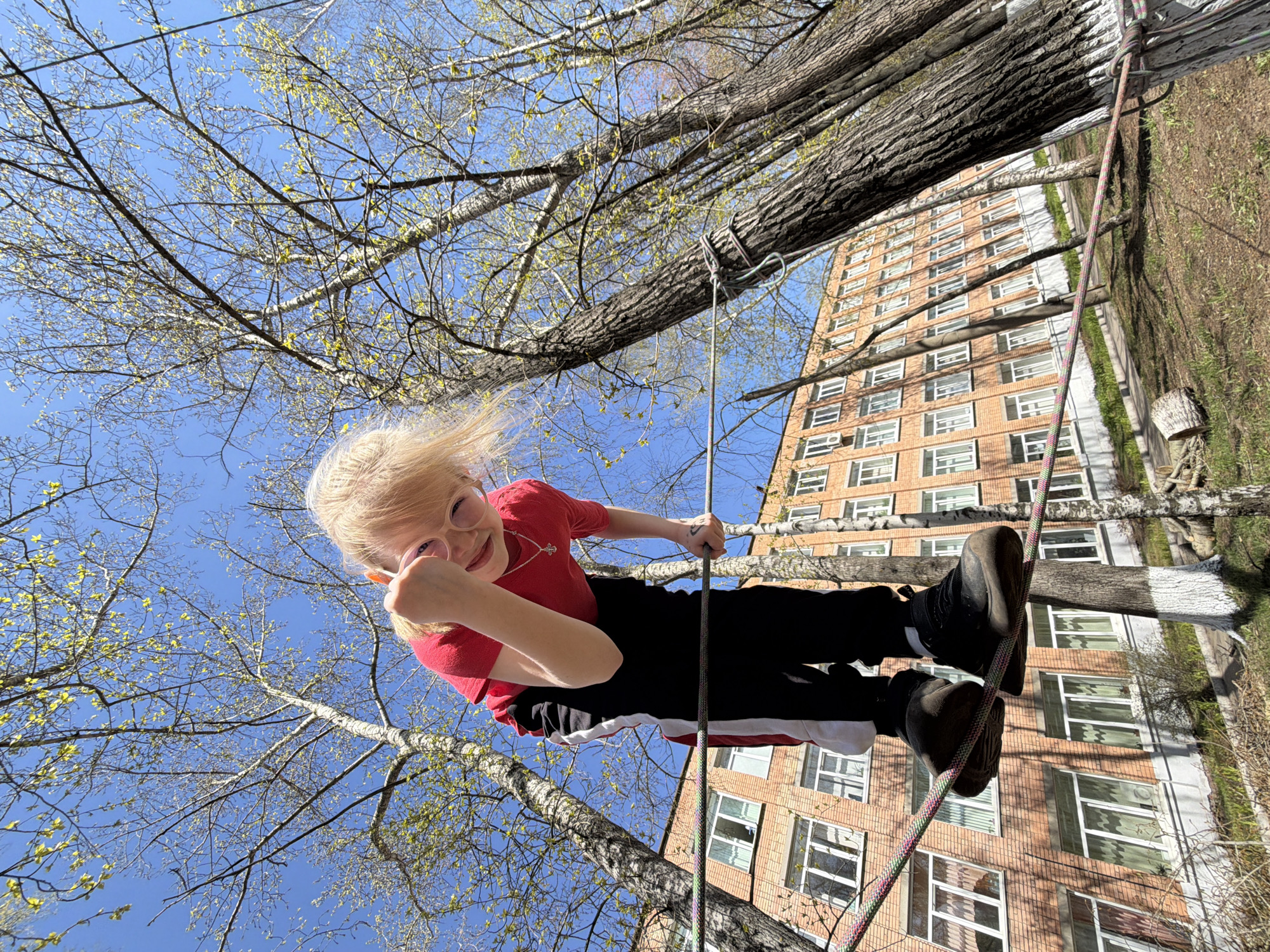 A child in a red t-shirt and black pants stands on a rope suspended between two trees. The child is smiling and holding onto a higher rope. In the background, a multi-story brick building is visible. The location is Izhevsk, Russia. The sky is blue with tree branches reaching towards it.