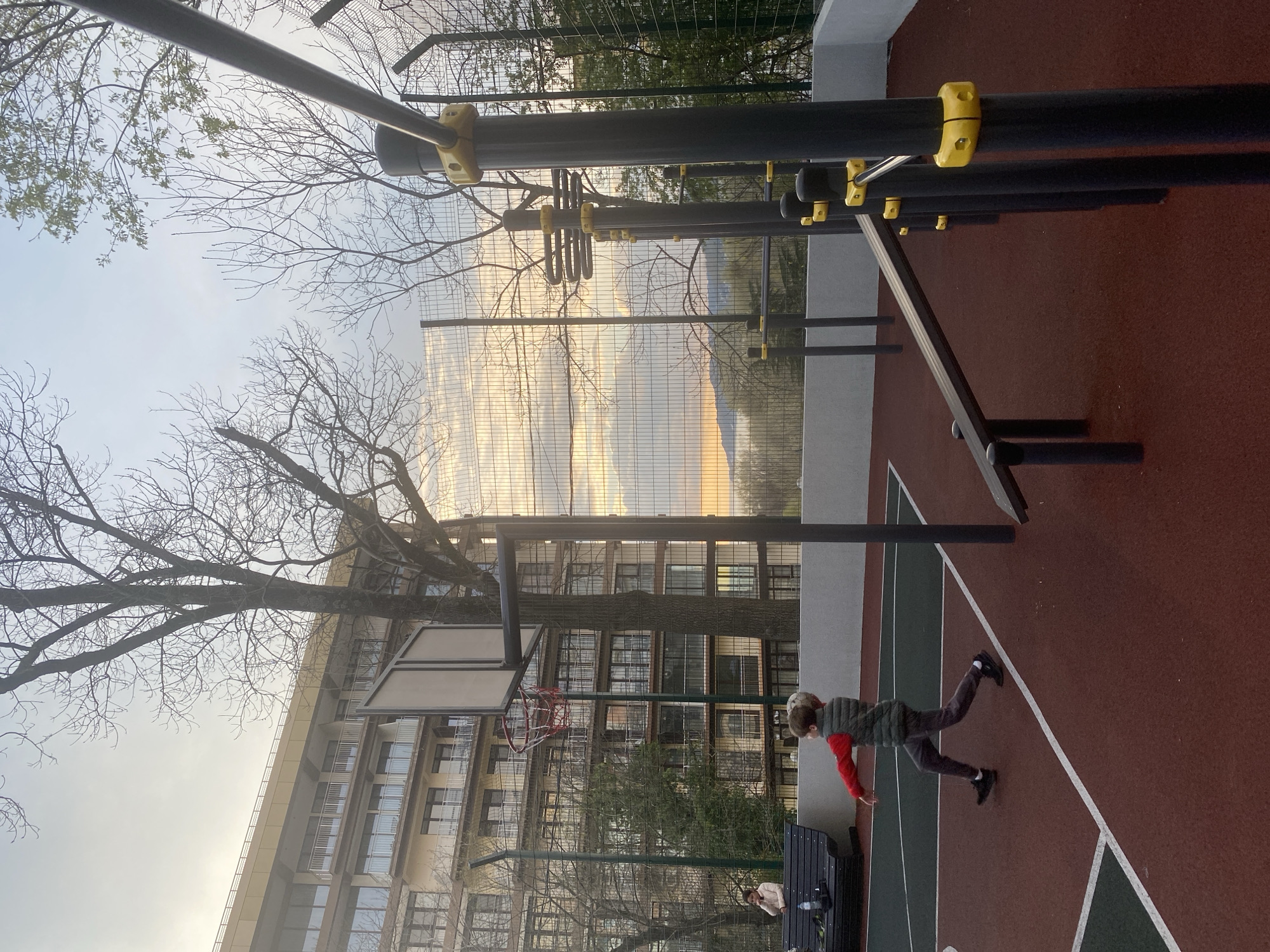 The image depicts an outdoor scene, possibly a sports facility, located in Sochi, Russia.
In the foreground, a young person is seen in motion on a basketball court, wearing dark pants, a red shirt, and a dark vest. The basketball court is marked with white lines and appears to be constructed with alternating sections of reddish-brown and green material.
Overhead, there is a complex structure of metal bars and supports, likely part of an exercise apparatus. The bars are predominantly black, with some yellow fixtures.
Behind the basketball court, a tall, multi-story building can be seen, with numerous windows and balconies. Adjacent to the building is a netted fence. Beyond the fence, a glimpse of a mountainous landscape and sky is visible. Bare tree branches fill the top of the frame, contrasting with the architecture and landscape.