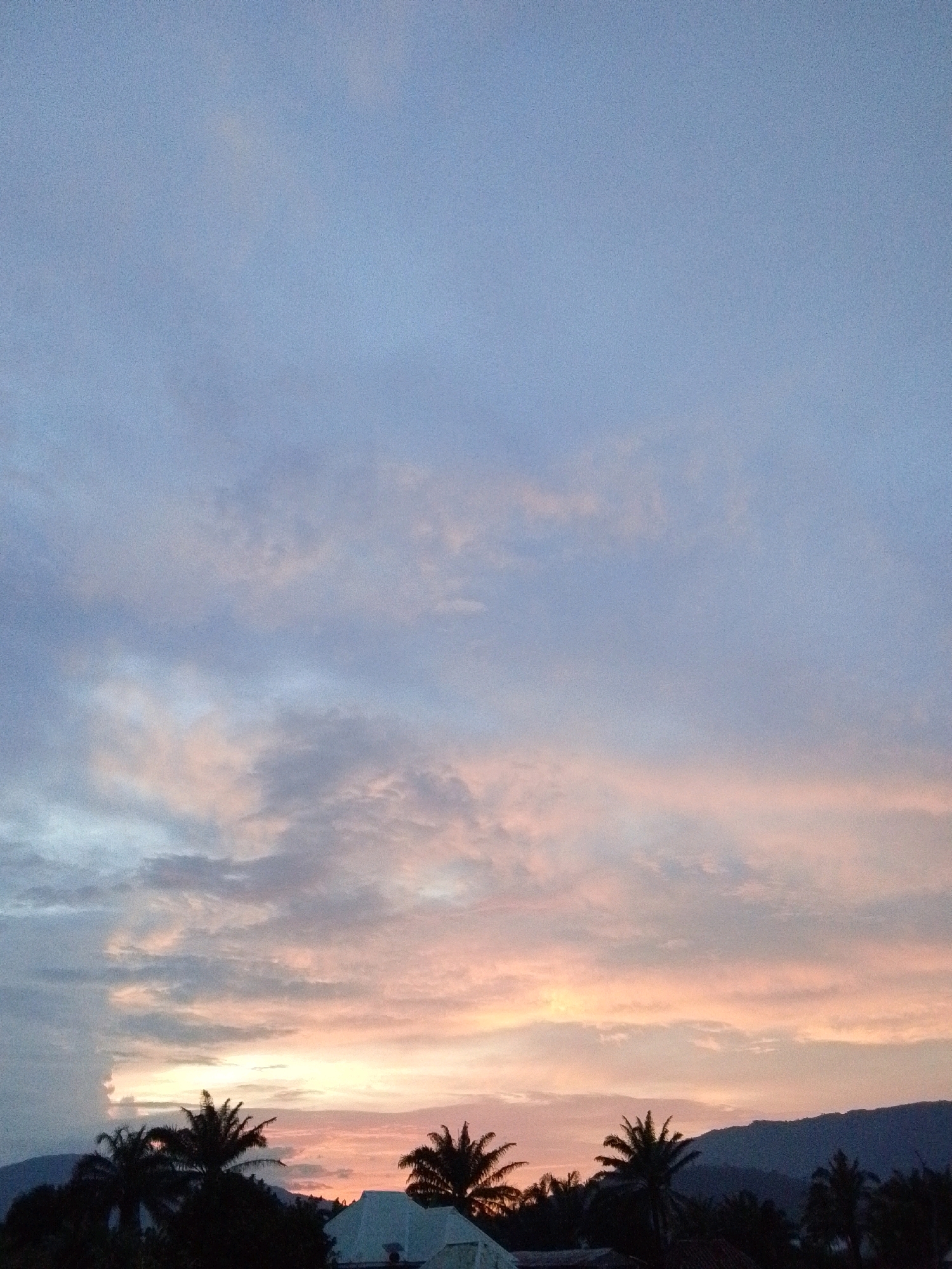This is a landscape photograph of a sunset over Serti, Nigeria. The sky is filled with soft hues of pink, orange, and blue, with scattered clouds. In the foreground, silhouetted palm trees and a residential building are visible. The overall impression is one of tranquility and natural beauty.
