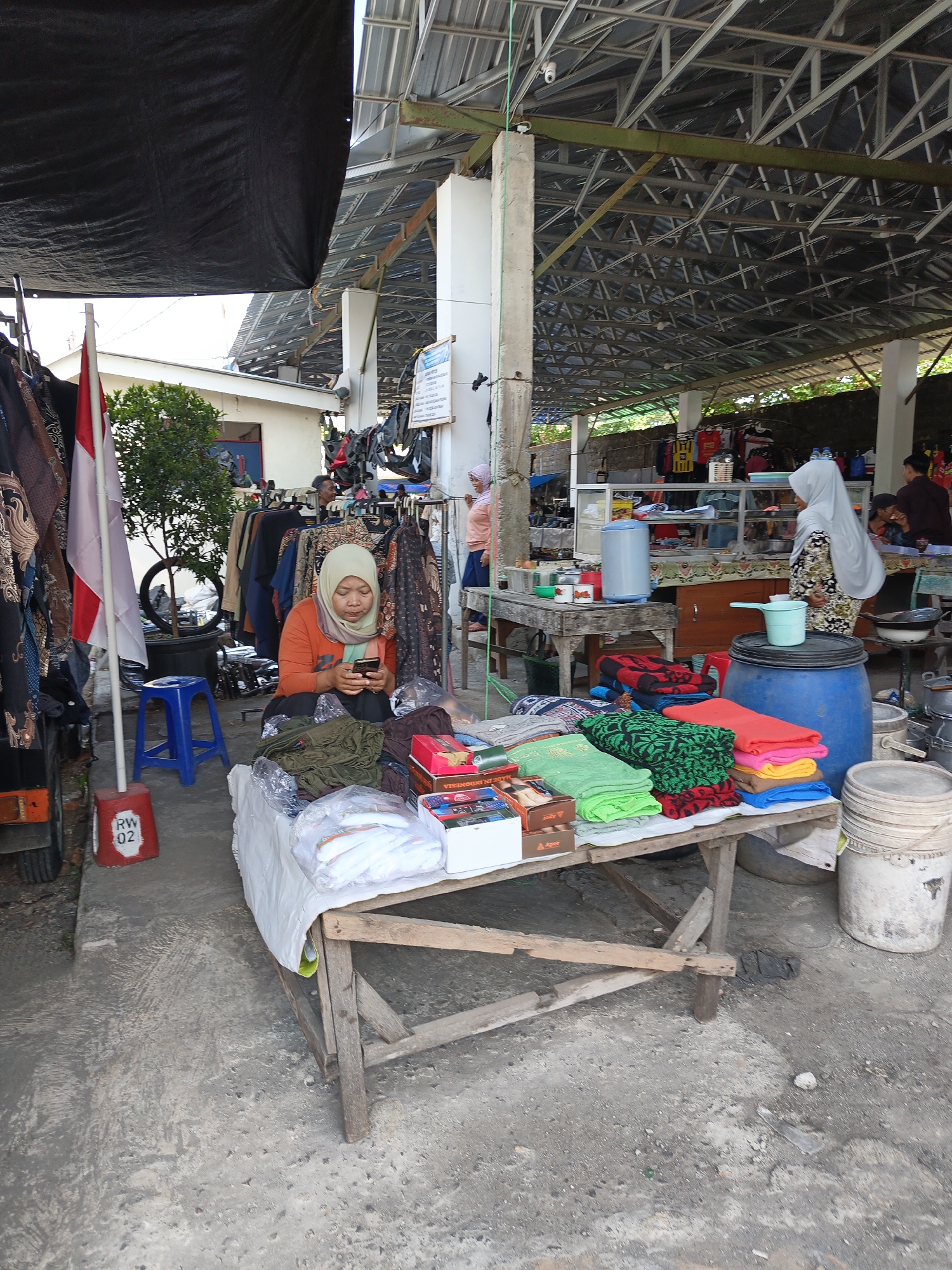 In Jatinom, Indonesia, a woman wearing an orange long-sleeved shirt and a hijab is seated at a stall, looking at her mobile phone. She is surrounded by various items for sale, including stacks of colorful folded fabrics, packaged goods, and clothing racks in the background. The stall is set up outdoors under a covered structure with a metal roof and concrete pillars. Nearby, a blue plastic stool and a red and white marker with "RW 02" are visible. The ground is paved with concrete, showing some wear and tear. In the distance, other market stalls and people can be observed, indicating a bustling marketplace environment. The scene is illuminated by natural daylight.