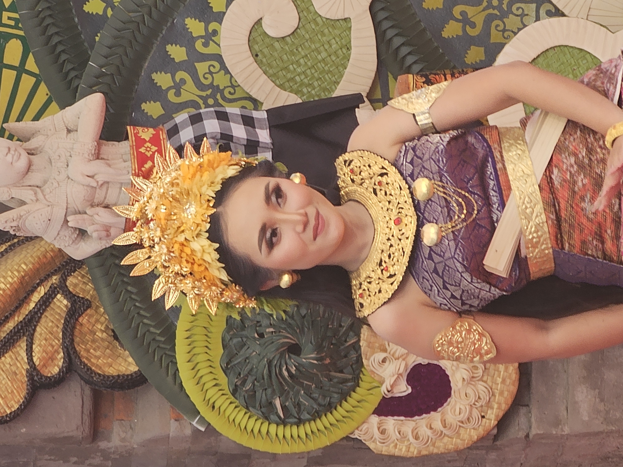 A young woman is posed in traditional Balinese attire, likely a dancer, with ornate golden headdress and jewelry, including a large collar and armbands. She wears a purple and gold patterned garment with a colorful woven skirt. Her makeup is dramatic, emphasizing her eyes. Behind her, elaborate decorations of woven palm leaves and a carved stone figure, possibly a deity or mythical being, are visible. The background includes a brick wall, suggesting an outdoor or temple setting. The overall impression is of a cultural performance or ceremony, possibly in a temple or public space in Denpasar, Indonesia, given the distinctive Balinese artistic elements.