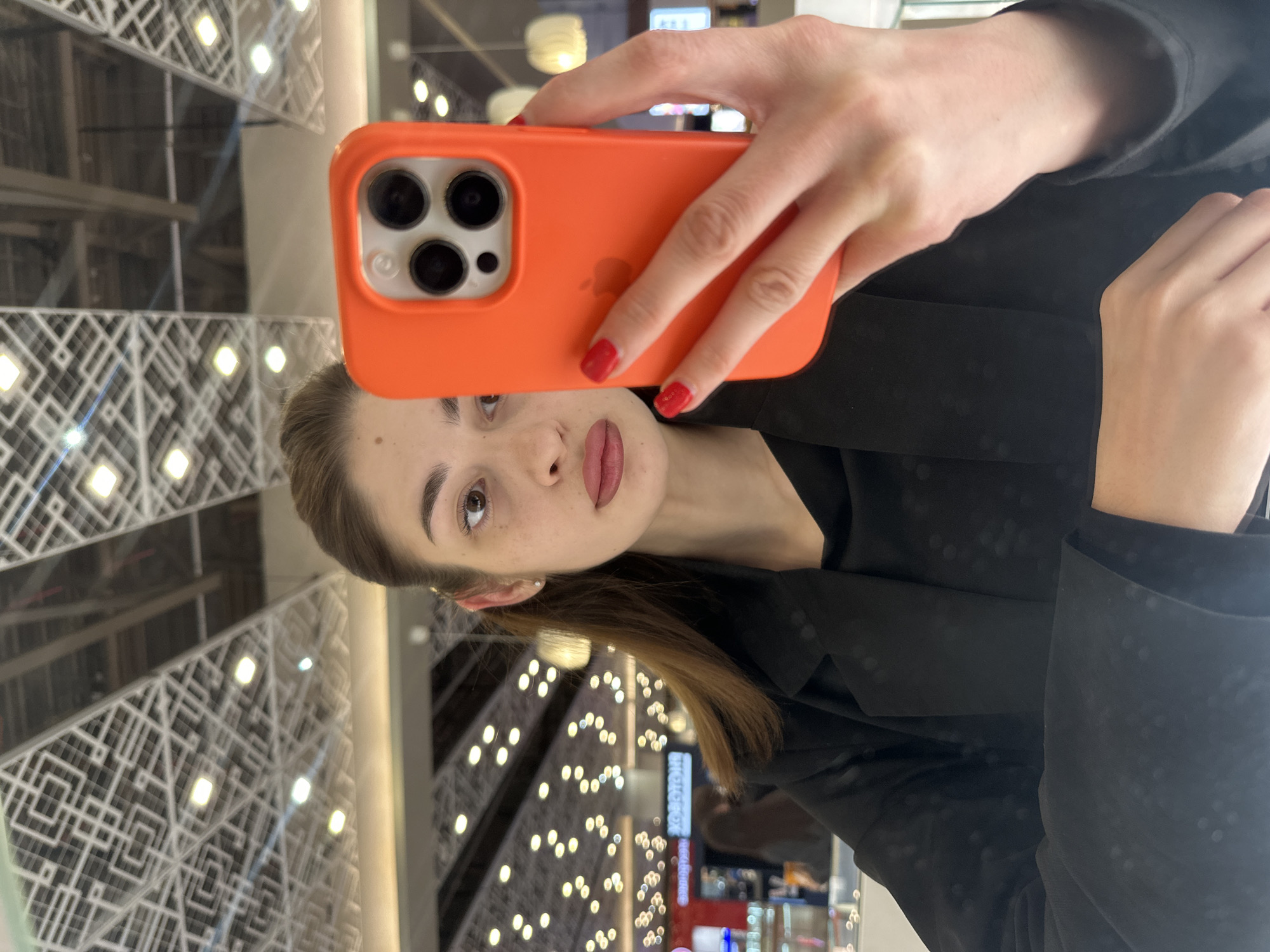 The image is a mirror selfie capturing a young adult female in the Greenwich Shopping Center, Yekaterinburg, Russia. The subject is depicted from the chest up, with her head slightly angled to her left. She has fair skin, brown eyes, and dark brown hair pulled back, revealing a small stud earring in her visible right ear. Freckles are visible across her nose and cheeks, and her lips bear a pinkish tint. She is dressed in a dark, likely black, blazer or jacket over a dark, subtly textured top. Her fingernails are painted bright red.
She is holding an orange-cased iPhone, identified as a Pro model by its triple camera array on the rear, in her left hand, positioned centrally and partially obscuring the lower half of her face. A faint Apple logo is discernible through the orange silicone case. Her right hand is partially visible beneath the phone, resting on an unseen surface.
The background is a reflection of the indoor commercial environment. The ceiling features a prominent, intricate geometric pattern of white lines on a darker, reflective surface, interspersed with numerous square, recessed light fixtures. Beyond this, the blurred background shows a multitude of smaller, warm-toned circular lights, creating a bokeh effect, consistent with a large indoor space. Visible signage in the reflection includes a Cyrillic sign that appears to read "YOKORU", indicative of a restaurant, and another blurred blue and white sign on the left, consistent with a mobile operator like "TELE2".