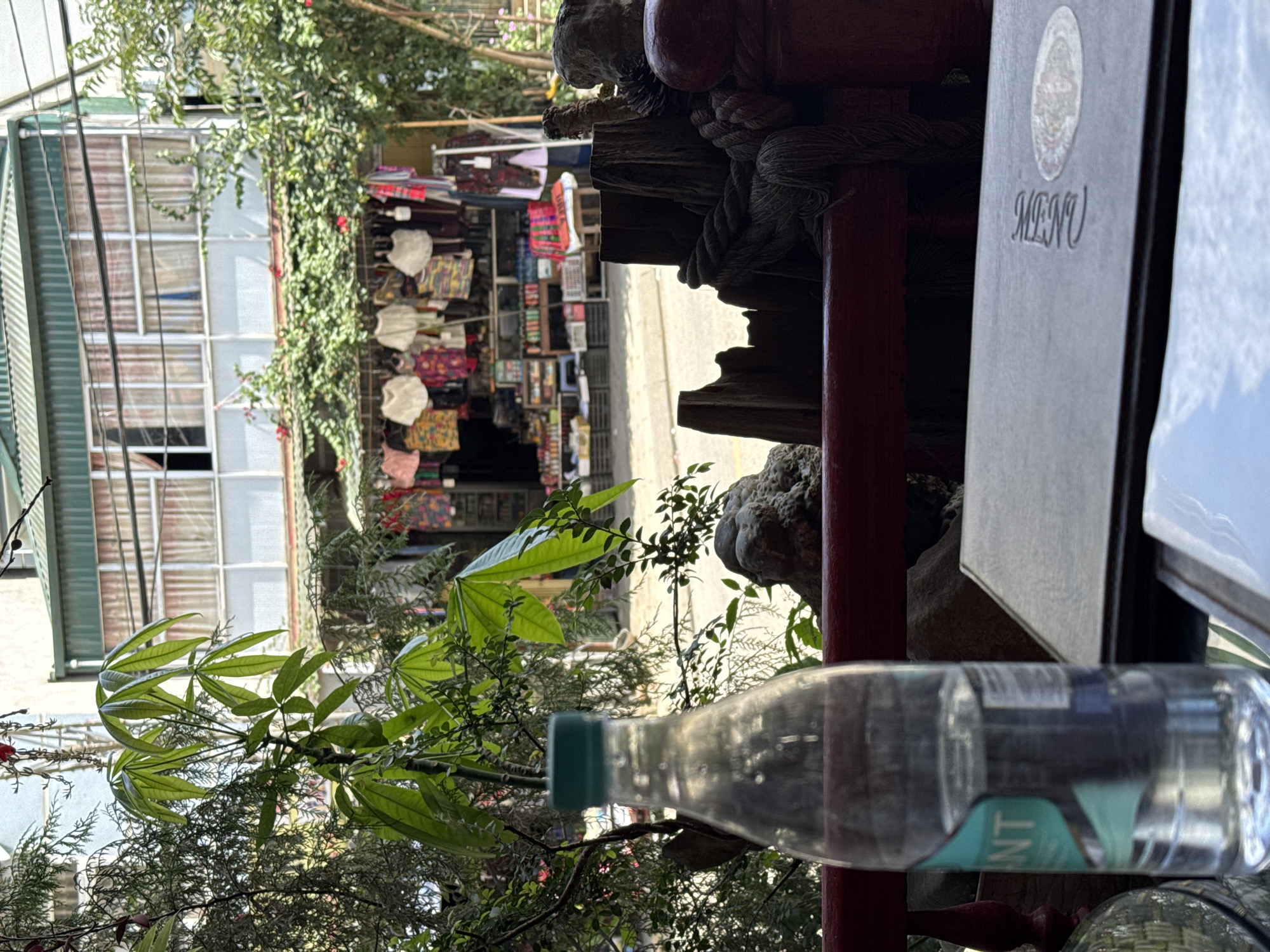 The photograph, taken in Đồng Văn Commune, Vietnam, shows an outdoor scene adjacent to a commercial district. A bottle of water is prominent in the foreground, positioned near a restaurant menu. The restaurant appears to be set up in an outdoor area decorated with plants. In the background, a building with numerous windows can be seen, along with a shop displaying merchandise, likely textiles or souvenirs. The scene has an ambient daylight, suggesting a midday or afternoon time. The angle of the shot and the elements included suggest it may have been taken while seated at the restaurant, capturing the view outwards.