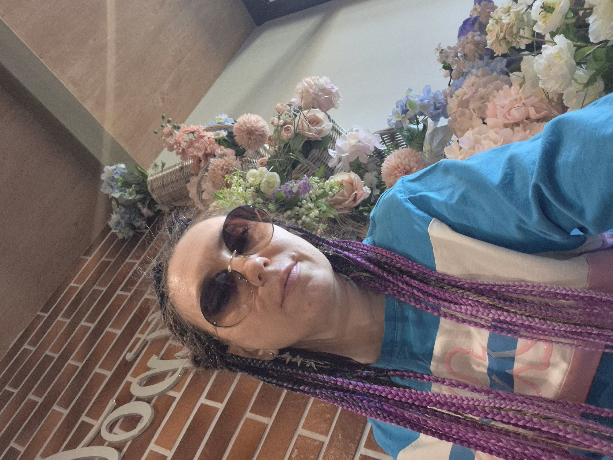 A woman with purple braids and sunglasses is centered in a close-up, slightly tilted shot. She is wearing a blue t-shirt with a white and pink design. Behind her is an arrangement of light-colored artificial flowers in baskets. To the left, a brick wall is partially visible with large, white lettering. Based on the visible features, the image may have been taken in Votkinsk, Russia, but this cannot be definitively determined without additional context.