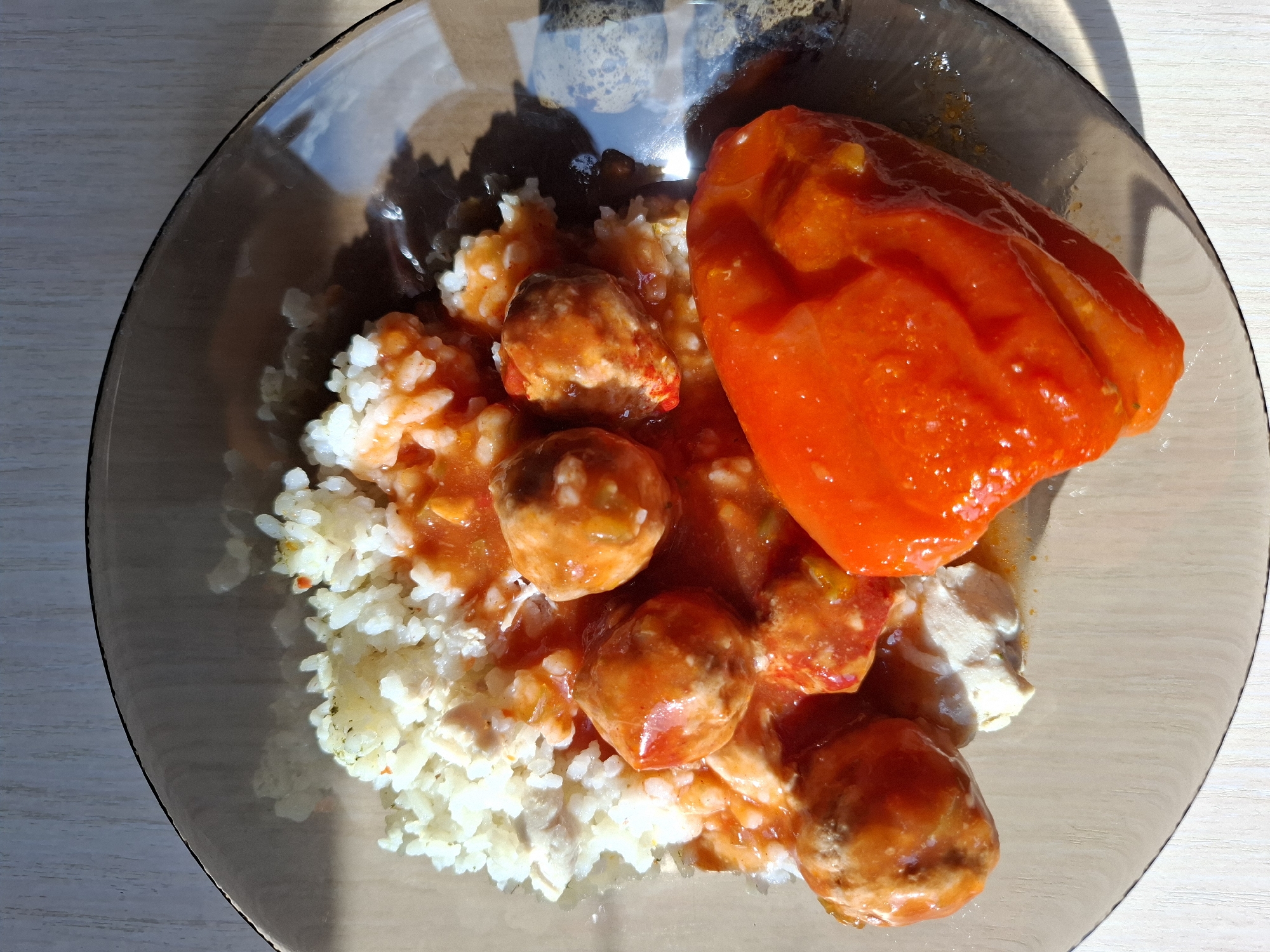 A close-up, overhead shot shows a portion of a meal on a clear glass plate. The plate rests on a light-colored, wood-grain surface. The meal consists of white rice topped with meatballs in a red sauce. There is also a large piece of red bell pepper, also covered in sauce, to the right of the meatballs. A small, pale portion of what appears to be a dairy product, possibly sour cream or yogurt, is visible beneath the meatballs. The lighting suggests natural light, casting shadows on the plate and the surface beneath. There is no information available to determine the location of this image in Tyumen, Russia.