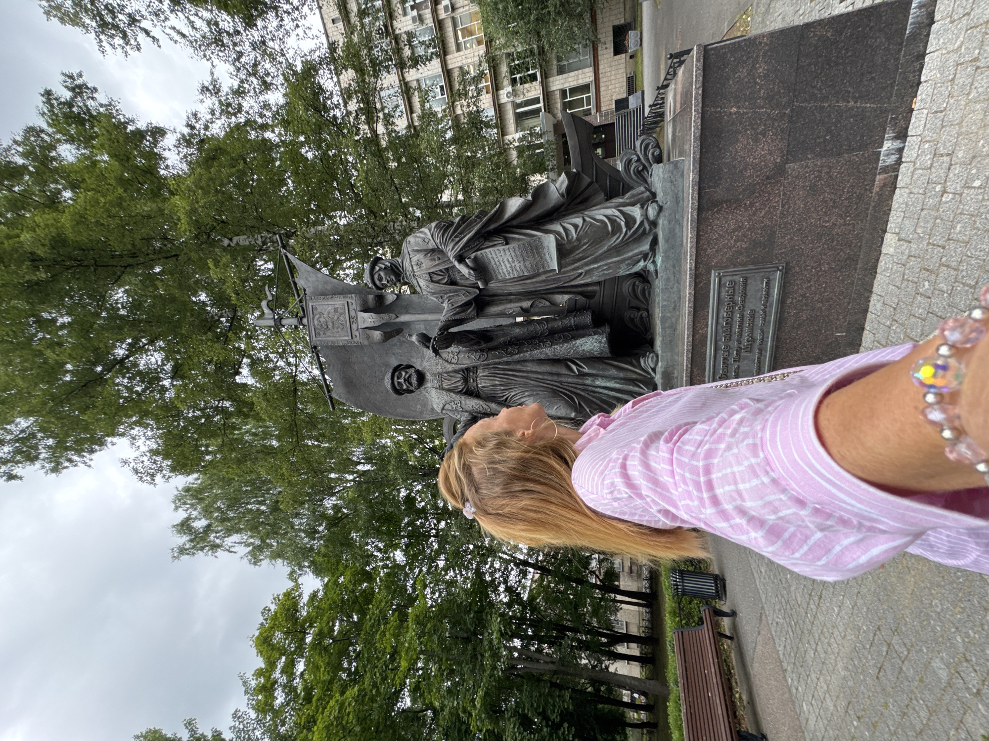 The image captures a woman from the chest down, facing away from the camera and looking towards a bronze monument. She has blonde hair with a pink scrunchie and is wearing a pink and white striped button-up shirt. She has a bracelet on her wrist with various beads, some of which are clear and faceted.
The monument is large and features two figures in historical attire, one holding a scroll and the other a bird. They stand on a base that appears to be made of dark, polished stone, with a plaque inscribed in Russian. Behind the monument, there are trees with green foliage and a building with multiple windows and air conditioning units. To the left of the monument, a wooden bench is visible along a paved walkway. The ground is a mix of cobblestones and paved sections. The sky is overcast.
The inscription on the plaque reads: "Святые Благоверные Князь Петр и Княгиня Феврония Муромские". This translates to "Holy Blessed Princes Peter and Fevronia of Murom". The location is Ladozhskoe Ozero, Russia.