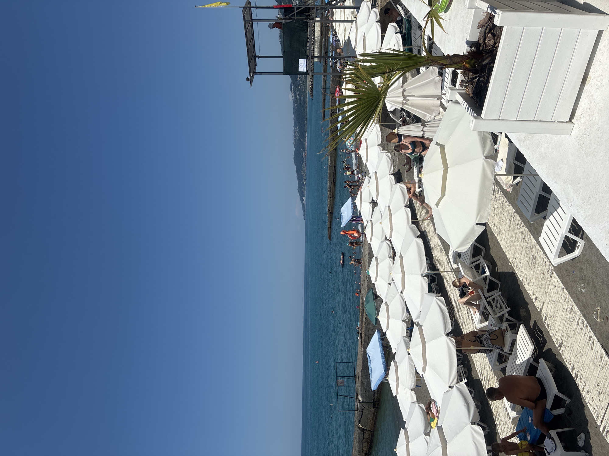 The image shows a crowded beach scene in Sochi, Russia on a sunny day. The beach is lined with white umbrellas and lounge chairs, and people are seen sunbathing, swimming, and relaxing. In the background, the Black Sea stretches to the horizon, with a mountainous coastline visible in the distance. A lifeguard tower stands on the right side of the image, with a person visible in the station. Some people are playing a card game on lounge chairs in the foreground. The overall scene depicts a typical summer day at a popular tourist destination.
