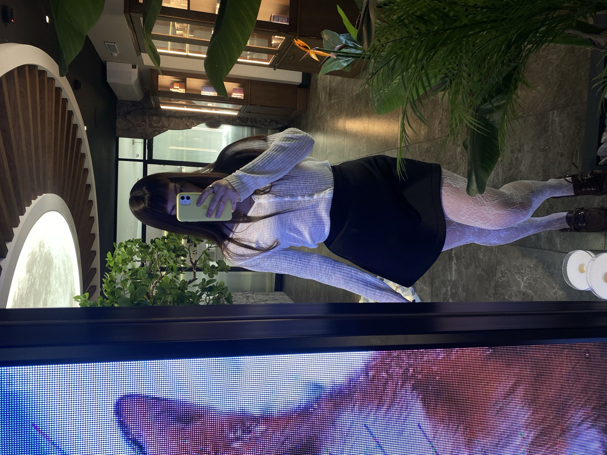 A person is taking a selfie in front of a large LED display, which is positioned in a room with indoor plants. The person is wearing a light-colored cardigan, a black skirt, and patterned tights, and brown boots. They are holding a cell phone, presumably to capture the photo. The location includes architectural elements, such as a wall with a distinctive design, and a glimpse of shelving with various objects. The plants add to the interior decor. The selfie is being taken in a mirror or reflective surface, creating a duplicated view of the scene. Without further evidence, it is impossible to confirm if the image was taken in Votkinsk, Russia.