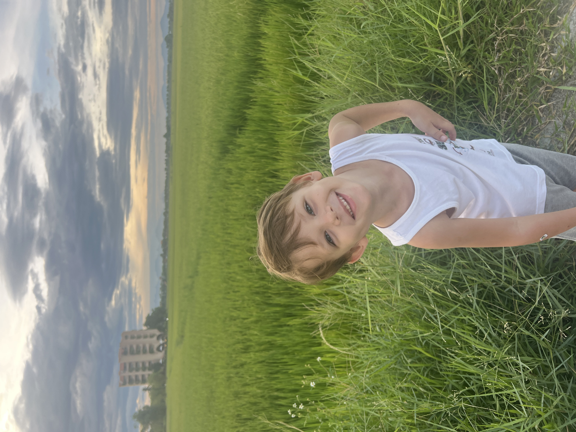 A young boy with light brown hair and blue eyes is pictured in a rice paddy field in Hoi An, Vietnam. He is wearing a white tank top and gray shorts, and he is smiling at the camera. The rice paddy is lush and green, and in the background, there is a building and mountains. The sky is cloudy, with some patches of blue and yellow. The sun appears to be setting or rising, casting a warm glow over the scene.
