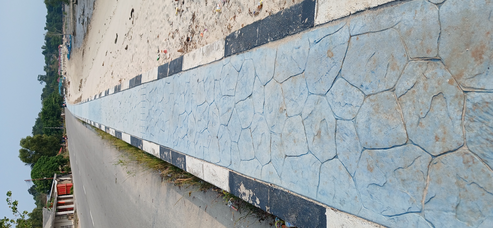 The image shows a concrete sidewalk with a blue stone pattern adjacent to a paved road. To the right of the sidewalk is a sandy beach area with scattered litter. In the background, there are trees and some buildings. Further down the road, several people can be seen on motorbikes, and there are small structures that appear to be beach huts. Boats are also visible on the water in the distance. The scene suggests a coastal area in Tanjung Balai Karimun, Indonesia.
