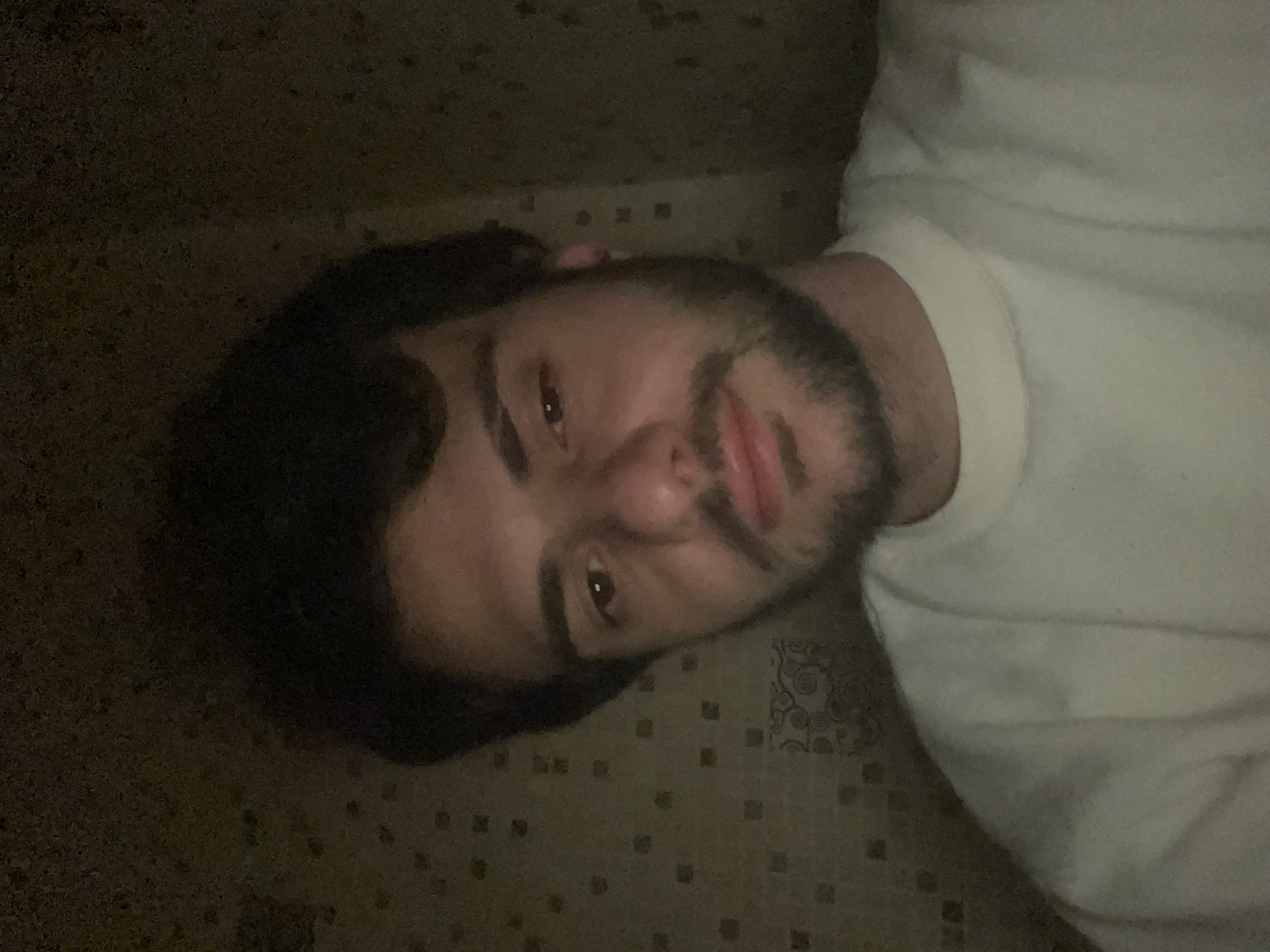 A young man with dark hair and a beard is depicted in a close-up, dimly lit selfie, taken as he lies down and looks up towards the camera. He is wearing a plain white t-shirt or top. His expression is neutral with a subtle smirk on his lips, and his brown eyes are directed at the viewer. A small pink object, possibly an earbud, is visible near his left ear.
The setting appears to be indoors. Behind his head, a dark, textured surface is visible, which could be a wall or a headboard. Beneath him, covering a significant portion of the frame, is a light brown or beige patterned fabric or surface, likely a bedspread or carpet. This surface is adorned with small, repeated square motifs and faint, larger swirling patterns. The lighting is low and artificial, suggesting the image was taken in the evening or at night. No specific visual elements within the image indicate the location as Tbilisi, Georgia. There is no visible text.