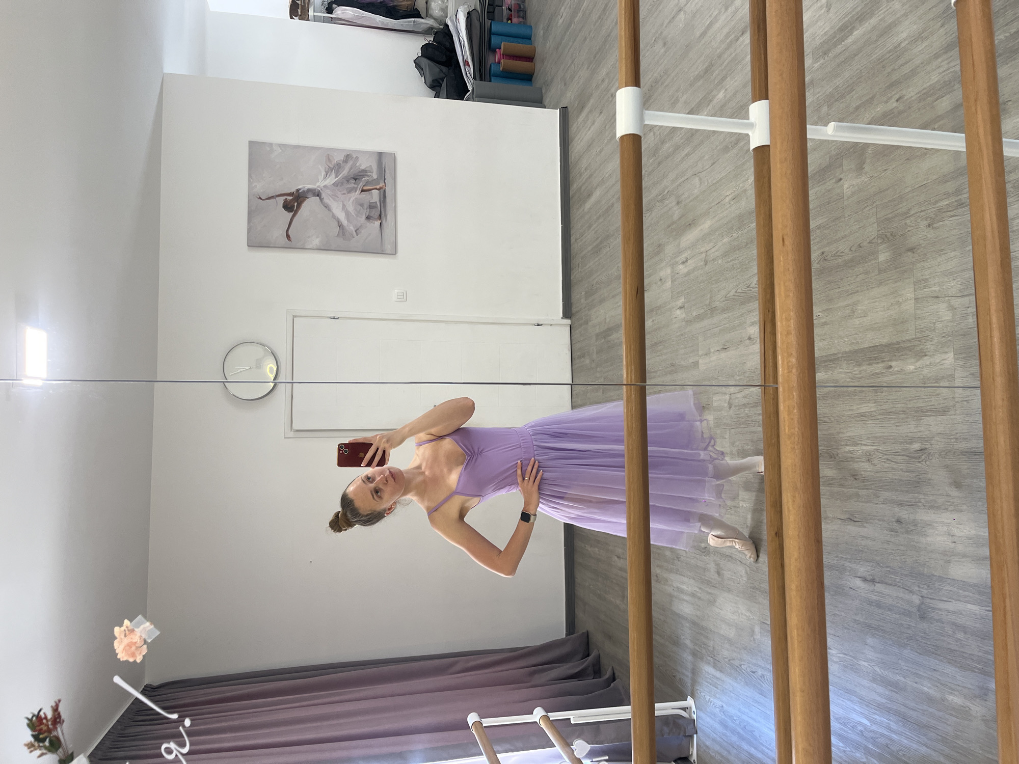 A woman in a lilac-colored dress and ballet slippers is taking a selfie in a mirror. She is standing in what appears to be a dance studio, as indicated by the ballet barre in the foreground and the painting of a ballerina on the wall. The floor is a light grey wood laminate. Various items are stored on a shelf in the upper left corner of the image. The city and specific location are Unknown, Unknown.