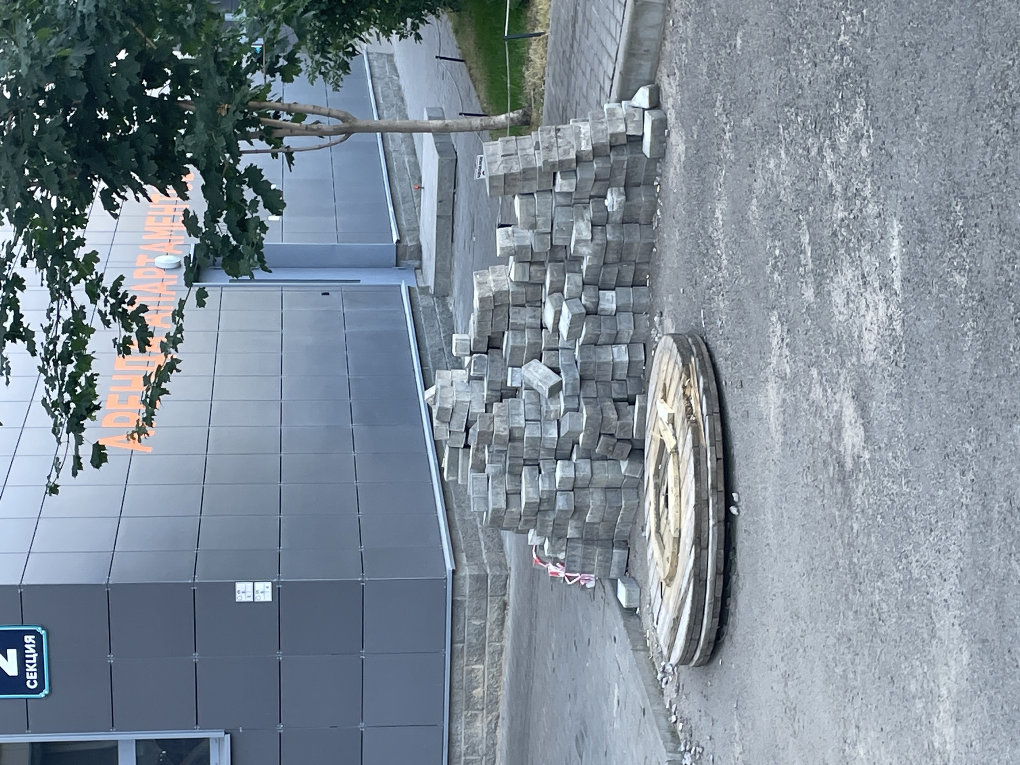 An outdoor, eye-level shot shows a construction site in front of a modern building in an unspecified Russian city. The building is clad in gray square tiles and features an orange sign with Russian text, partially obscured by a tree. To the left, a blue sign reads "2 СЕКЦИЯ" (2 SECTION).
In the foreground, a large pile of gray paving stones is stacked haphazardly. Adjacent to the stones is a large, circular wooden spool with a flat top, resting on the asphalt. The ground is a mix of asphalt and paved walkways. A small tree with green leaves grows to the right of the building. The scene appears to be under natural daylight.