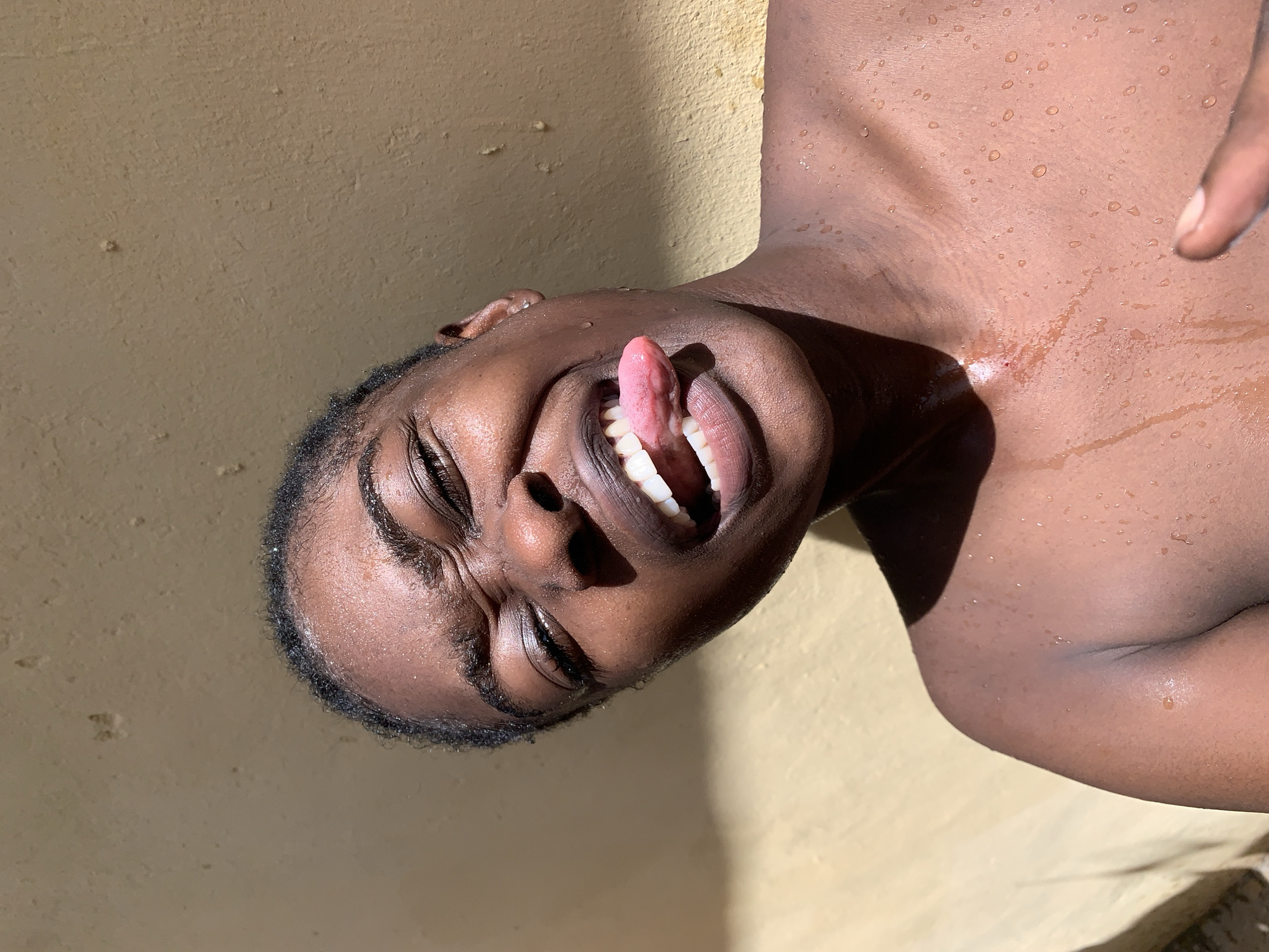 A dark-skinned individual with wet skin lies on their side on a tan-colored surface, possibly concrete or a wall. They are partially nude, with water droplets visible on their skin. Their eyes are closed, and their mouth is open, with their tongue extended. The background is a plain tan color. The location is Rafin Par, Nigeria. The time of day appears to be bright and sunny due to the distinct shadows.