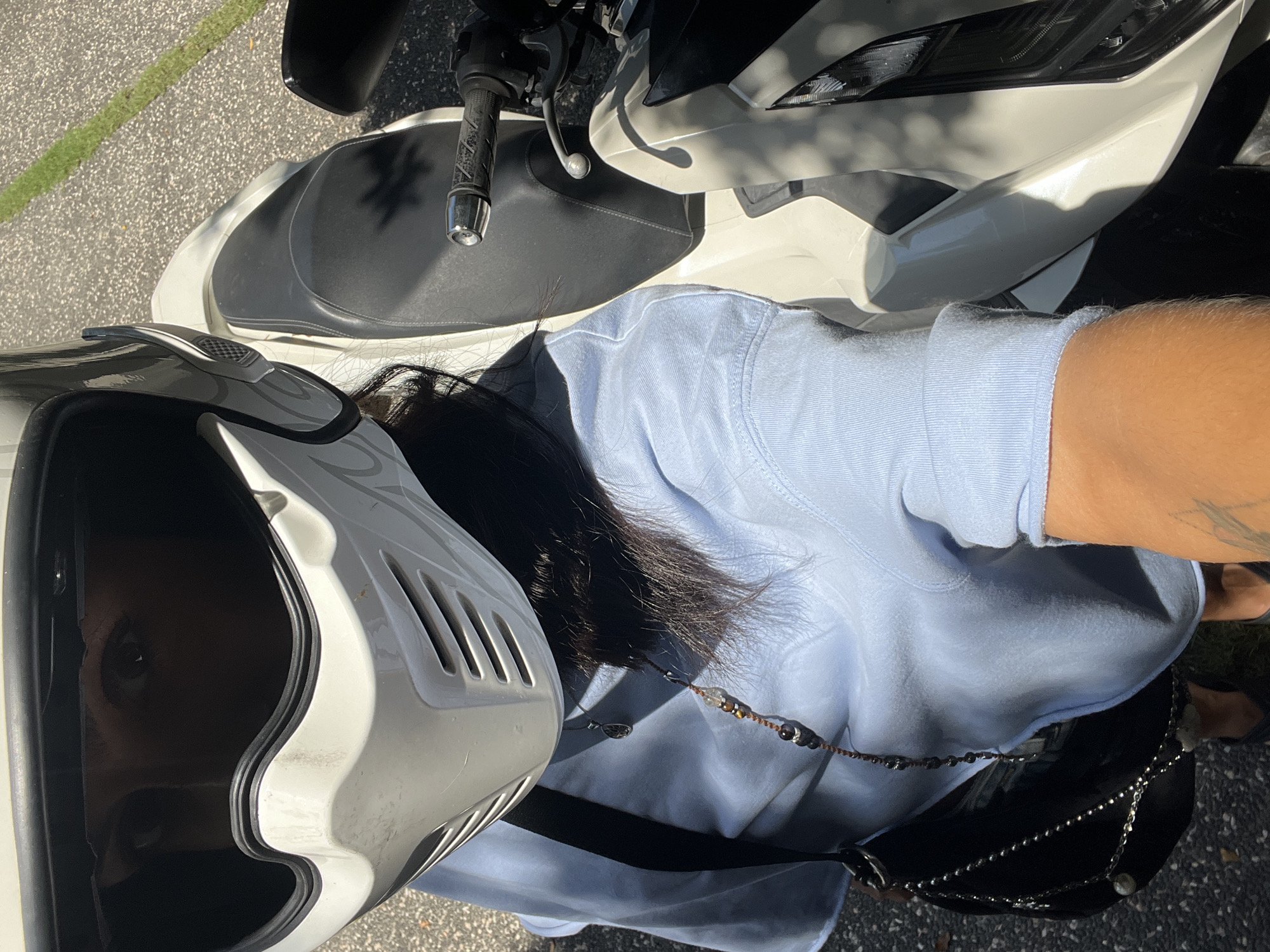 A person wearing a white helmet is sitting on a white scooter on asphalt. The person is wearing a light blue long-sleeved shirt and a black bag. The person has black hair. The location is not discernible from the image.