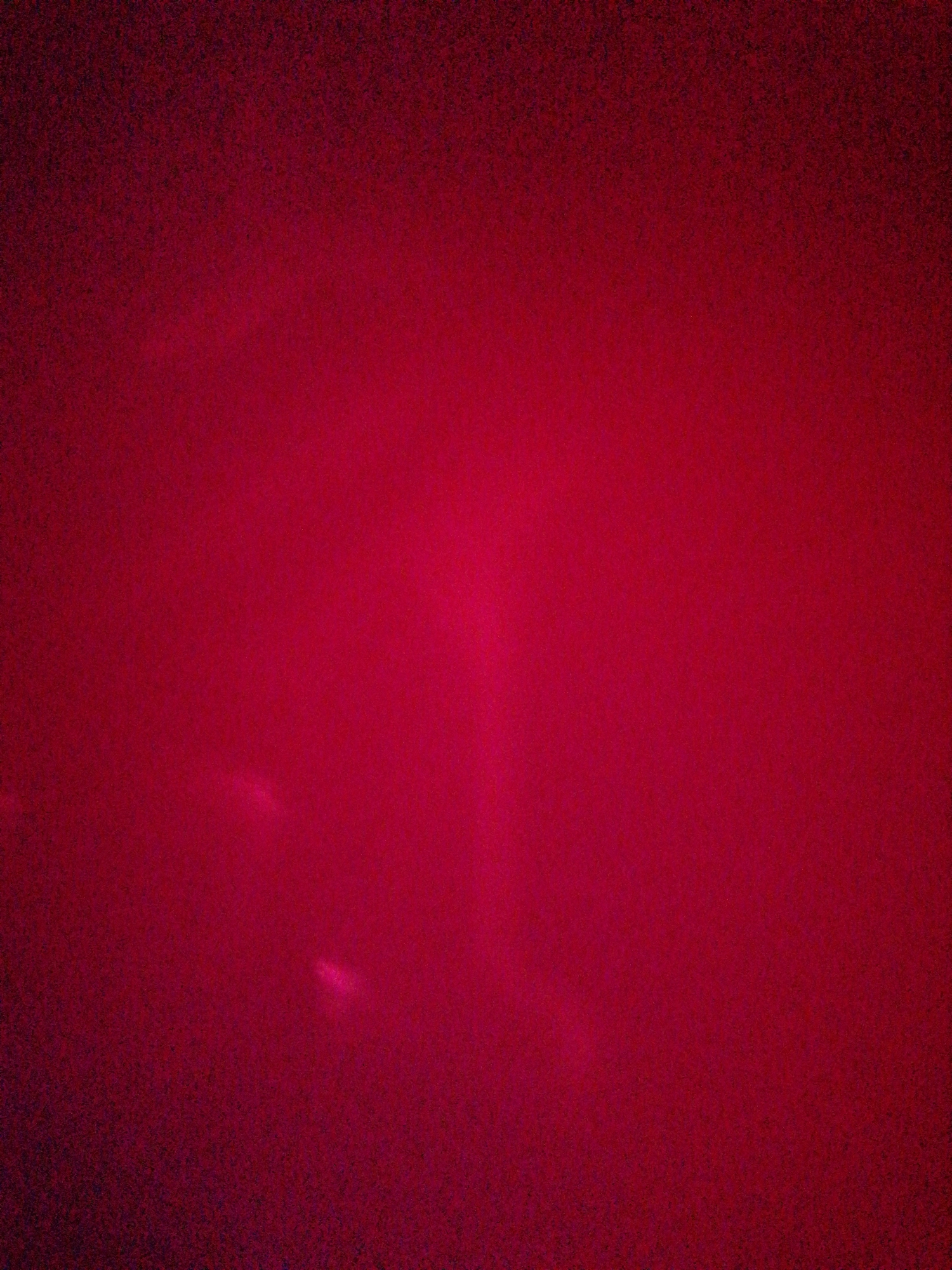 The image is a close-up, abstract view of a dark red, textured surface. The lighting is dim and uneven, creating a gradient of color from a deep, almost black red in the upper left corner to a brighter, more vibrant red in the lower right. There are faint, blurry streaks of light or highlights visible in the lower left quadrant. The texture of the surface appears to be slightly grainy or fibrous. The image provides no discernible information about location, people, or specific objects.