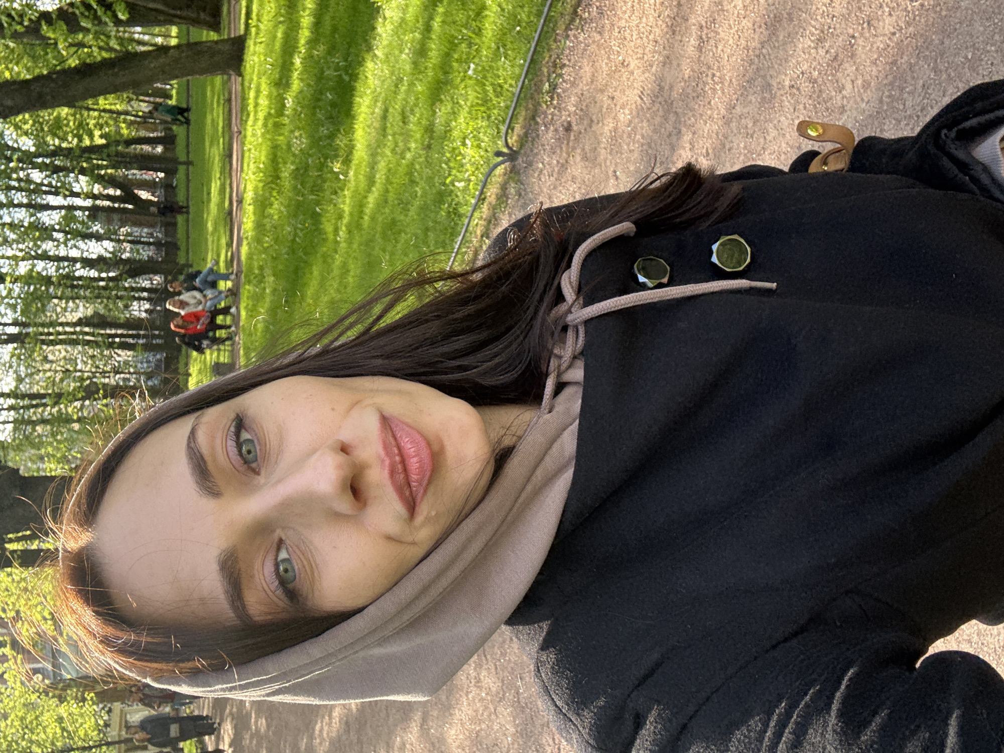 The image is a selfie taken by a woman with fair skin, dark brown hair, and green eyes. She is wearing a black coat with a beige hoodie underneath. The background shows a park setting, likely in Saint Petersburg, Russia, given the presence of lush green lawns, trees, and people sitting on benches. A gravel path is visible.