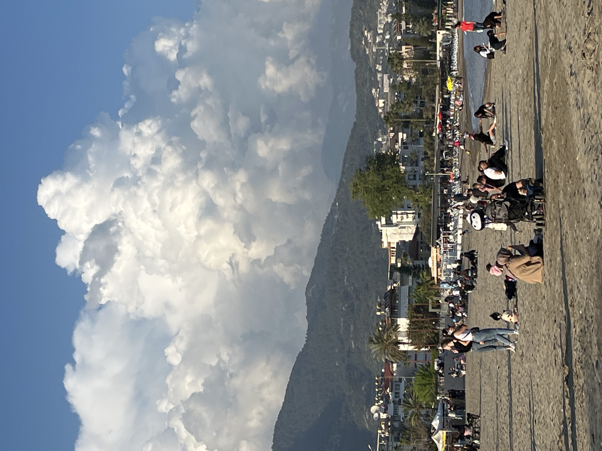 The image depicts a beach scene in Alanya, Turkey. A wide beach is adjacent to a paved promenade with numerous people walking and relaxing. The beach is sandy, with people near the water's edge. Buildings line the promenade. A large mountain rises behind the buildings, partially obscured by clouds. The sky is blue with large, puffy cumulus clouds. Palm trees and streetlights are visible along the promenade.