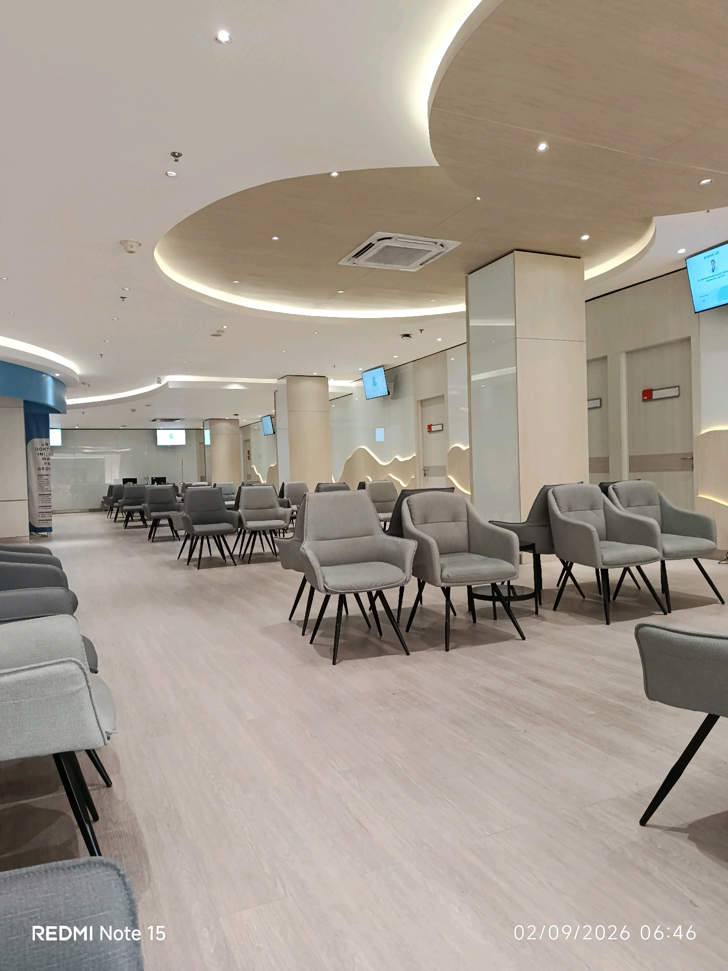 The image depicts the interior of a modern, well-lit waiting area, likely a hospital or clinic, in Waru, Indonesia. The space is furnished with numerous rows of empty, grey upholstered chairs with black legs, arranged on a light-colored wooden floor. The ceiling features recessed lighting and a curved, elevated section. Several tall, cream-colored pillars are present, and some walls have decorative wavy patterns. Large digital screens are mounted on the walls, displaying information or advertisements. There are also closed doors visible, each with a small red sign. The overall impression is one of clean, contemporary design and an organized, spacious environment. No people are visible in the frame. The timestamp in the lower right corner indicates the image was taken on February 9, 2026, at 06:46. The text "REDMI Note 15" is also visible in the lower left corner, indicating the device used to capture the image.