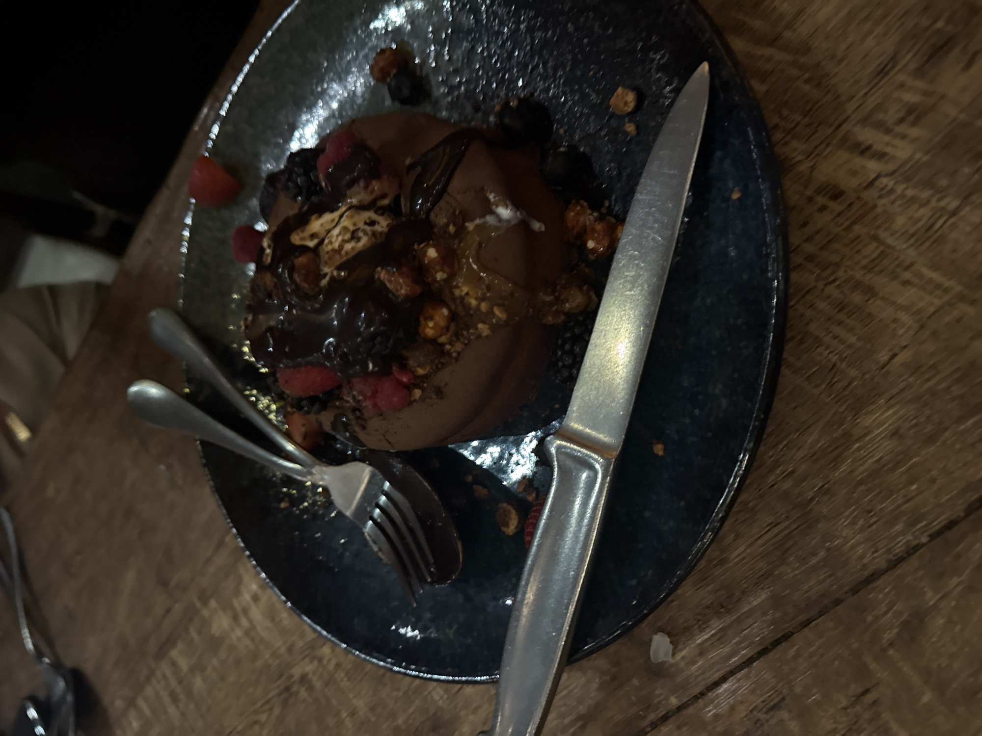 On a wooden table, a dark speckled plate holds a dessert consisting of a chocolate-colored base, topped with a dark sauce, red berries, dark berries, and toasted bits. A silver fork and spoon rest near the edge of the plate, partially covered by the dessert. A silver knife lies to the right of the dessert. The image may have been taken at a restaurant or cafe in Mexico City, Mexico.