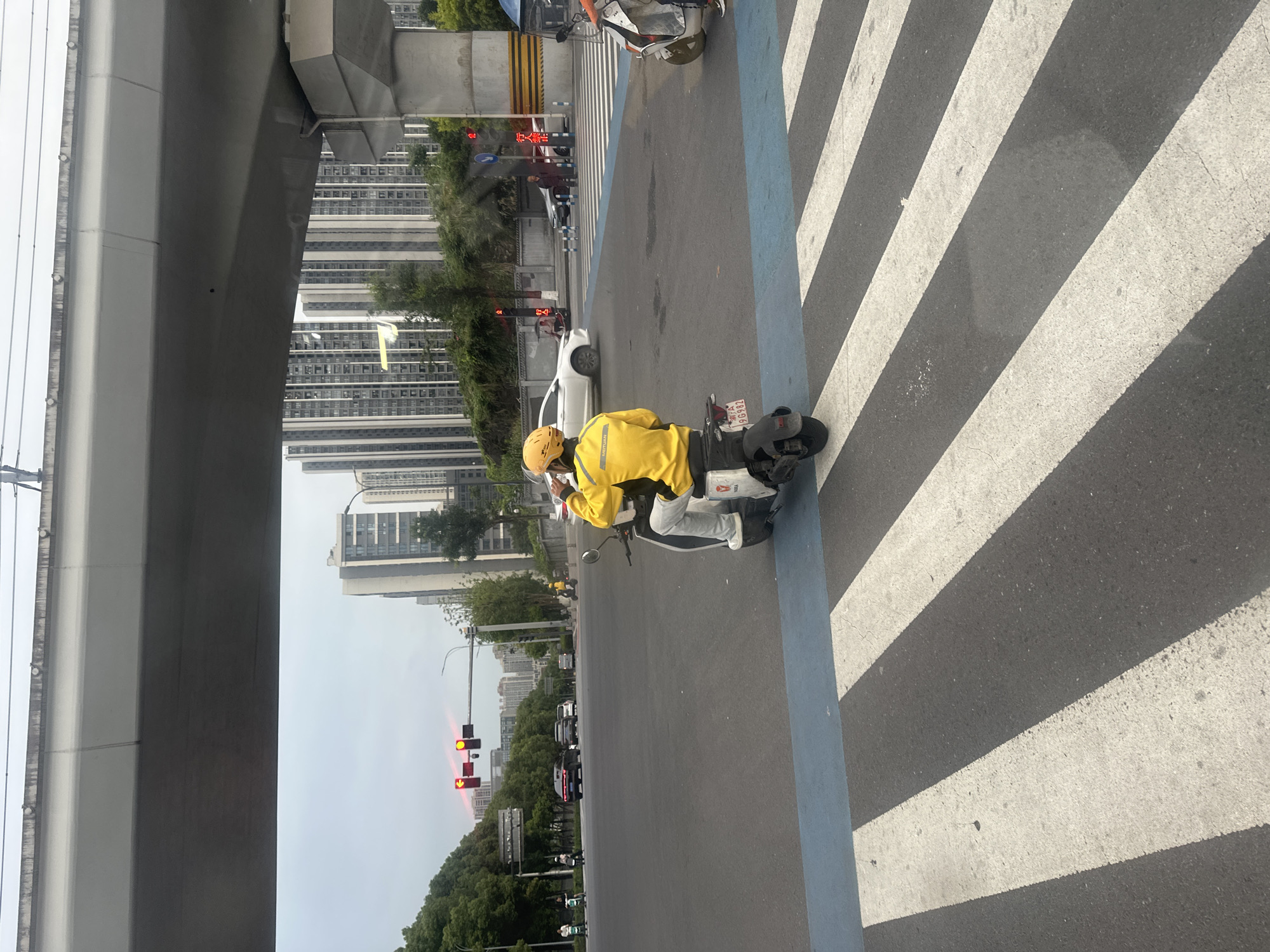 An overhead shot captures a street scene in the Chongqing High-tech Zone, China, characterized by a prominent elevated roadway and surrounding urban architecture. In the foreground, a person wearing a bright yellow jacket and helmet rides a white electric scooter, positioned on a pedestrian crossing marked with bold white stripes and a blue stripe. The scooter has a license plate with Chinese characters and numbers. Behind the scooterist, a white car drives through an intersection, with traffic lights showing red. Across the street, tall, modern residential buildings dominate the skyline, interspersed with trees. Other vehicles, including cars and smaller electric vehicles, are visible in the background, navigating the intersection under the grey, overcast sky. A concrete pillar supporting the elevated roadway is also visible on the right side of the frame.