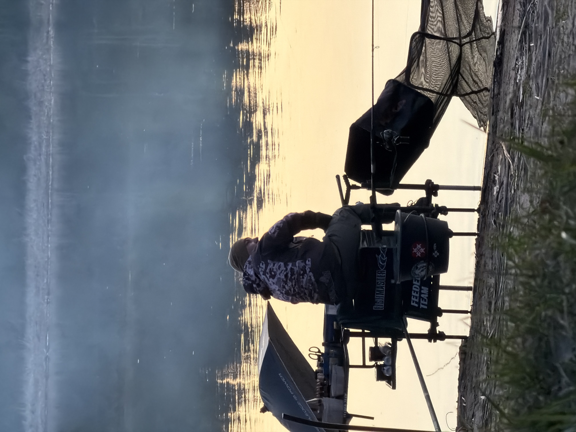 A person is seated on a fishing platform next to a body of water, likely a lake or river. The platform is equipped with fishing gear, including a keepnet, feeder, and umbrella. The person is wearing a patterned jacket and a hat. The body of water reflects the sky, which appears to be during either sunrise or sunset. The location is likely near the city of Chaykovsky, Russia, due to the type of fishing gear and environment shown in the image. Grass and earth are visible on the edge of the bank. The side of the platform visible reads “FISHMASTER FEEDER TEAM”.
