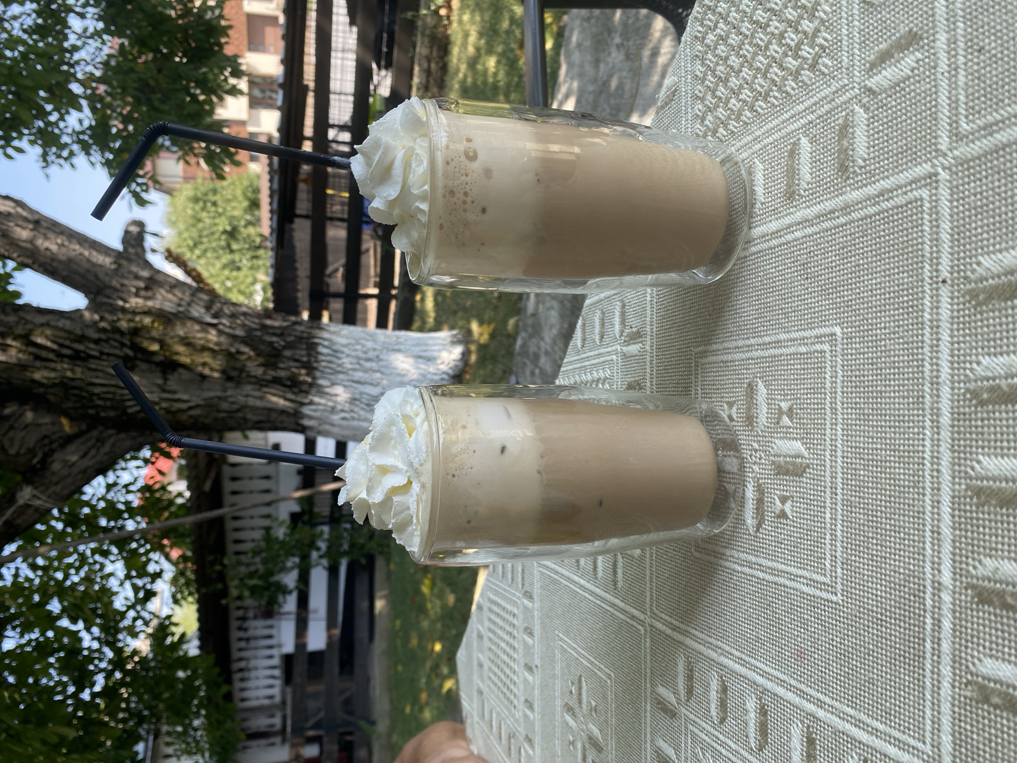 Two tall glasses filled with iced coffee topped with whipped cream and black straws are positioned on a light-colored, textured tablecloth. The glasses are double-walled, indicating they are likely insulated. In the background, a large tree trunk with white paint accents stands prominently. Beyond the tree, a wooden fence encloses a yard with green grass. In the further background, buildings with visible roofs and windows suggest a residential or commercial area, possibly within Târgu Neamț, Romania, given the context. The scene is outdoors, under natural daylight, with foliage from trees visible.