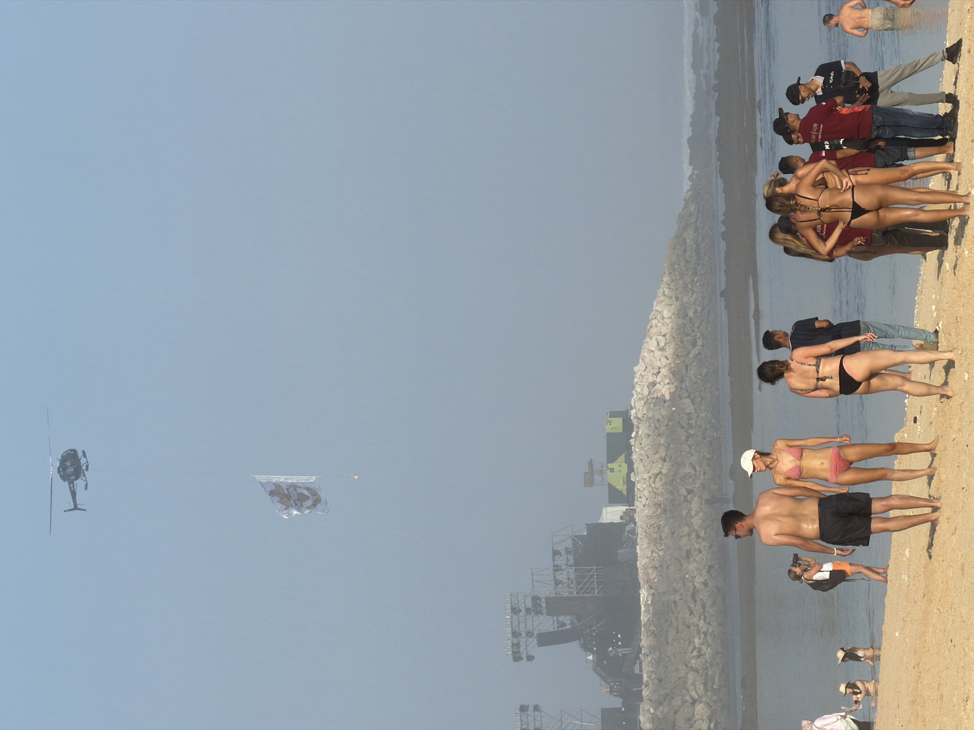 A helicopter flies in the sky above a beach with a flag attached to it. In the background, a large stage structure with lighting equipment is visible. On the beach, several people are standing and interacting, some in swimwear and others in casual clothing. A few individuals are in the shallow water. The environment appears to be a sunny day with a clear sky, although there is a slight haze.