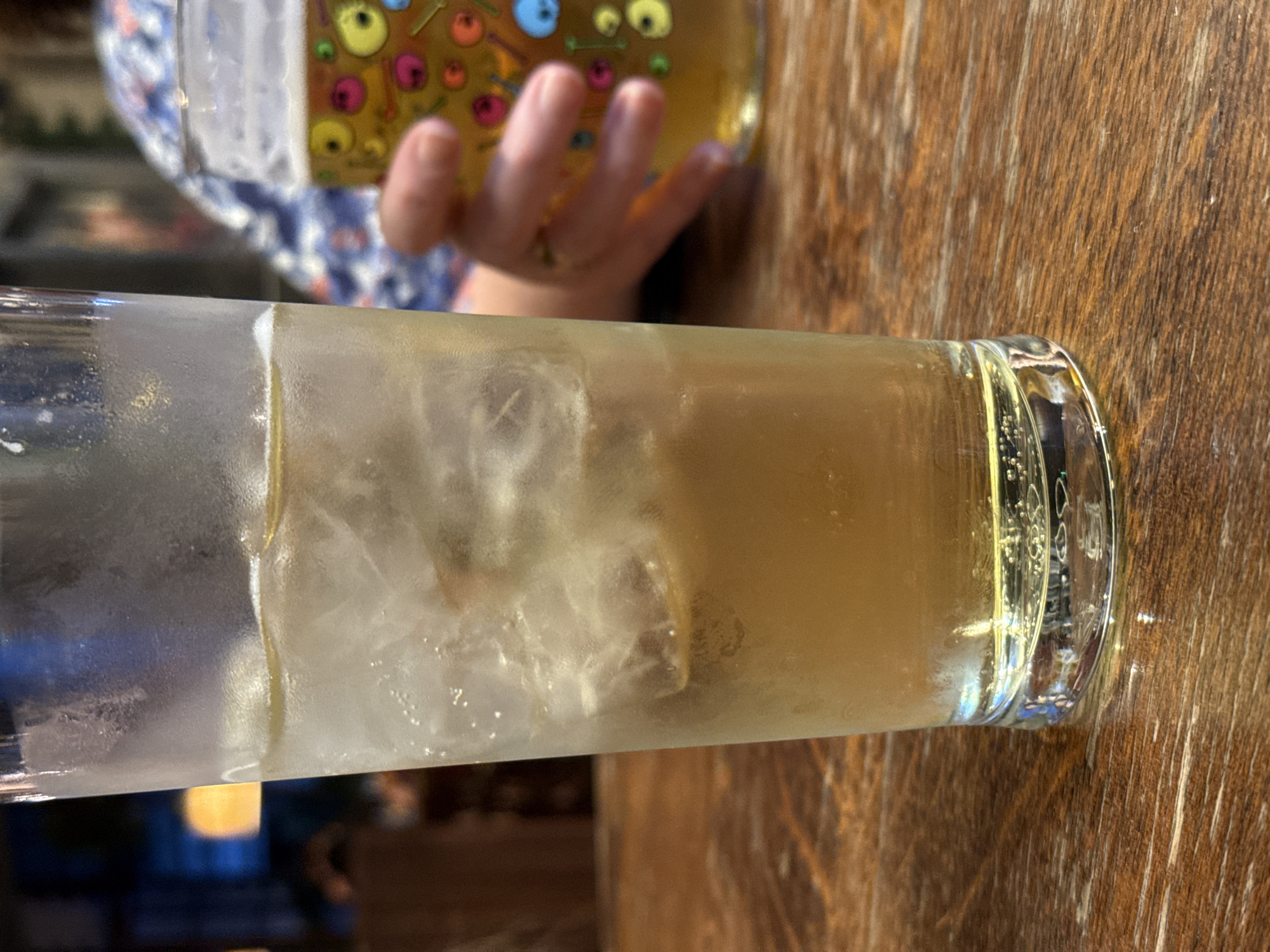 A horizontal close-up shows a tall glass filled with ice and a light brown liquid, likely a cocktail, resting on a wooden table. Condensation is visible on the outside of the glass. In the background, a child's hand reaches for a bottle with colorful cartoon-like illustrations. The surrounding environment appears to be a bar or restaurant, with a warm, dimly lit atmosphere, however, the specific location in London, United Kingdom cannot be determined from the provided information.