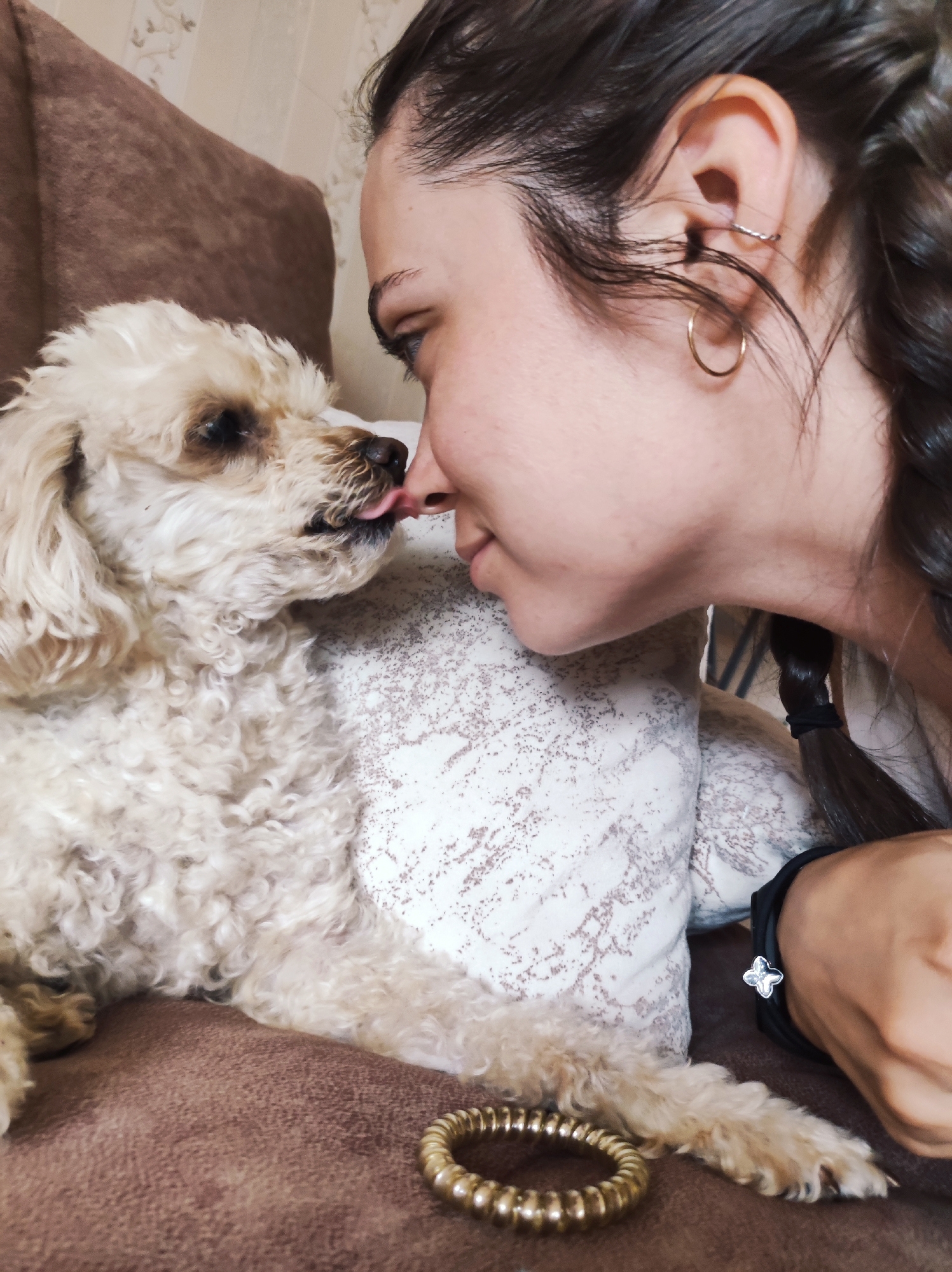 A young woman with dark hair in a braid is leaning in to kiss a small, light-colored poodle on the nose. The dog’s tongue is sticking out, licking the woman's nose. They are both on a brown sofa. The woman is wearing a gold hoop earring and a bracelet with a clover charm. A second gold bracelet is on the sofa near the dog's paw. The background shows a patterned pillow and a wall with a floral wallpaper. The scene is an indoor residential setting in Chaikovsky, Russia.