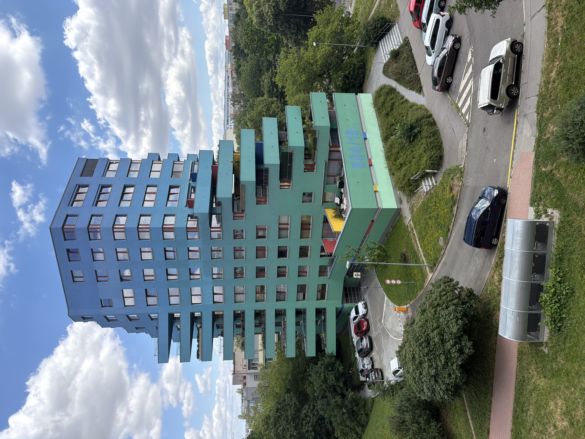 This image captures a modern, multi-story apartment building with a distinctive stepped design, featuring protruding balconies and a facade that transitions from blue at the top to teal and then bright green at the base. The building is located in Prague, Czechia. A parking lot with several cars, including a silver SUV, a dark sedan, and a white hatchback, is situated in front of the building. Adjacent to the parking lot is a grassy area with trees and shrubs. A paved walkway leads to the building's entrance, and a glass-covered structure, possibly a bike rack or storage unit, is visible in the foreground. The sky is blue with scattered white clouds, and the overall scene suggests a residential urban environment during daytime.