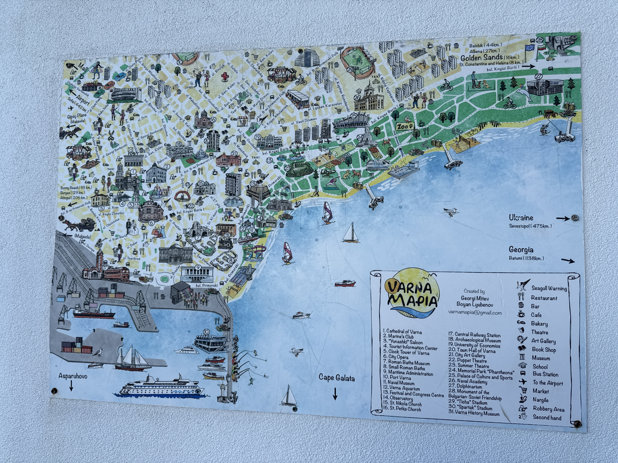 This image is a photograph of a stylized, hand-drawn map of Varna, Bulgaria, affixed to a textured, light-colored wall. The map is colorful and depicts various landmarks, streets, and the coastline.
The map itself is divided into several sections, with a prominent title "VARNA MAPIA" in the lower right quadrant. A legend is provided, listing numbered points of interest such as "Cathedral of Varna," "Marine's Club," "Zoo," and "Golden Sands." The map also indicates distances to other locations like "Balchik," "Albena," "Sunny Beach," and "Sofia." The sea is depicted on the right side of the map, with illustrations of boats, ships, and a lighthouse. The Asparuhovo bridge is labeled at the bottom left.
The wall on which the map is displayed appears to be outdoors, possibly a building exterior, given the texture and the way the light falls on it. The map is secured to the wall with visible metal fasteners at the bottom corners. The overall impression is that of an informational display in a public space.