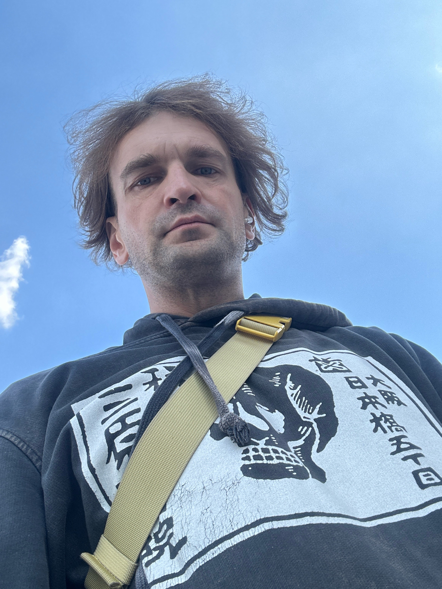 A low-angle, full-body selfie of a fair-skinned man with medium-length brown hair and blue eyes, looking up at the camera. He is wearing a dark hooded sweatshirt with a graphic print of a skull and Japanese characters. A light brown strap of a bag is visible across his chest. The background is a clear blue sky with a few wispy clouds, indicating an outdoor setting. The image does not contain any specific landmarks or details that would indicate a location in the city of Biysk, Russia.
