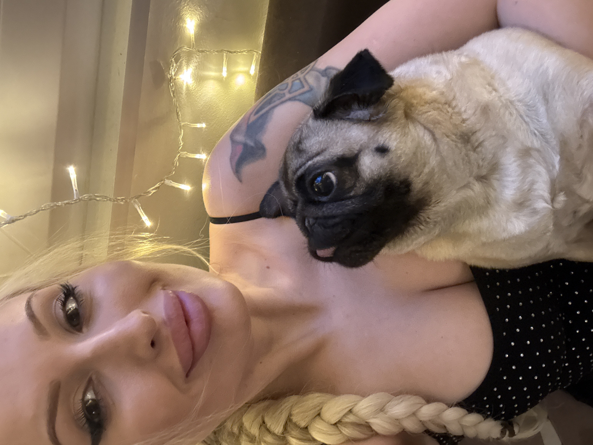 A woman with blonde hair in a braid, prominent makeup, and a black bejeweled top is holding a pug. The pug is looking upwards. They are indoors, with string lights visible in the background.