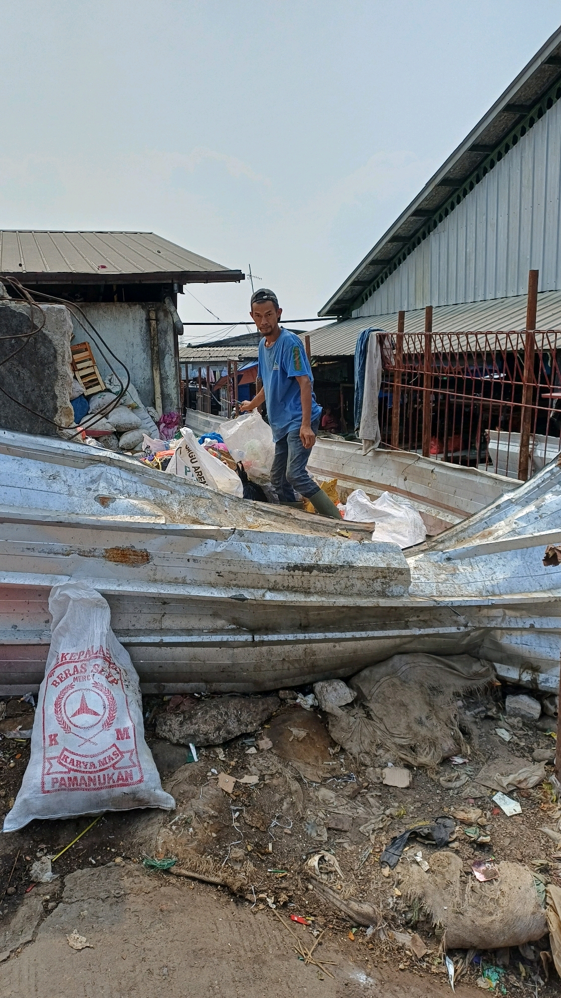 In Citeureup, Indonesia, a man in a blue t-shirt and jeans, wearing a cap and green boots, stands on a pile of corrugated metal sheets. He is holding a white plastic bag filled with trash. The scene appears to be a makeshift dump or recycling area, with more bags of trash and debris scattered around. In the background, there are several buildings with corrugated metal roofs, and a fence. The sky is clear and blue, indicating a sunny day.