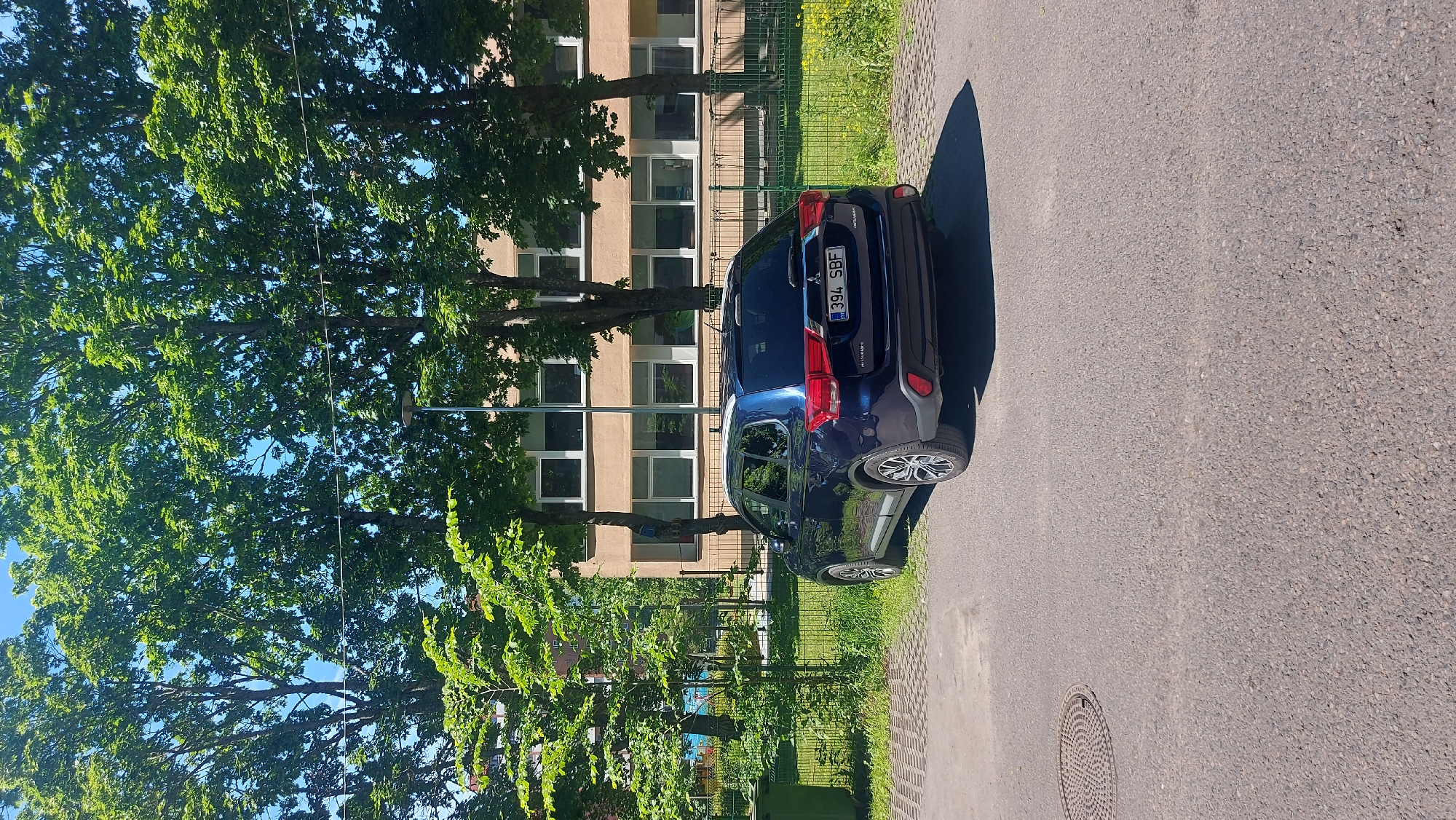 A dark blue Mitsubishi Outlander SUV is parked on an asphalt surface, adjacent to a grassy area and a building with a beige facade and white-framed windows. The vehicle has a license plate that reads "394 SBF". In the background, several large trees with lush green foliage dominate the scene, casting shadows. To the left of the vehicle, a green garbage bin is visible behind a wire fence. The sky is clear and blue. The location is the city of Tallinn, Estonia.