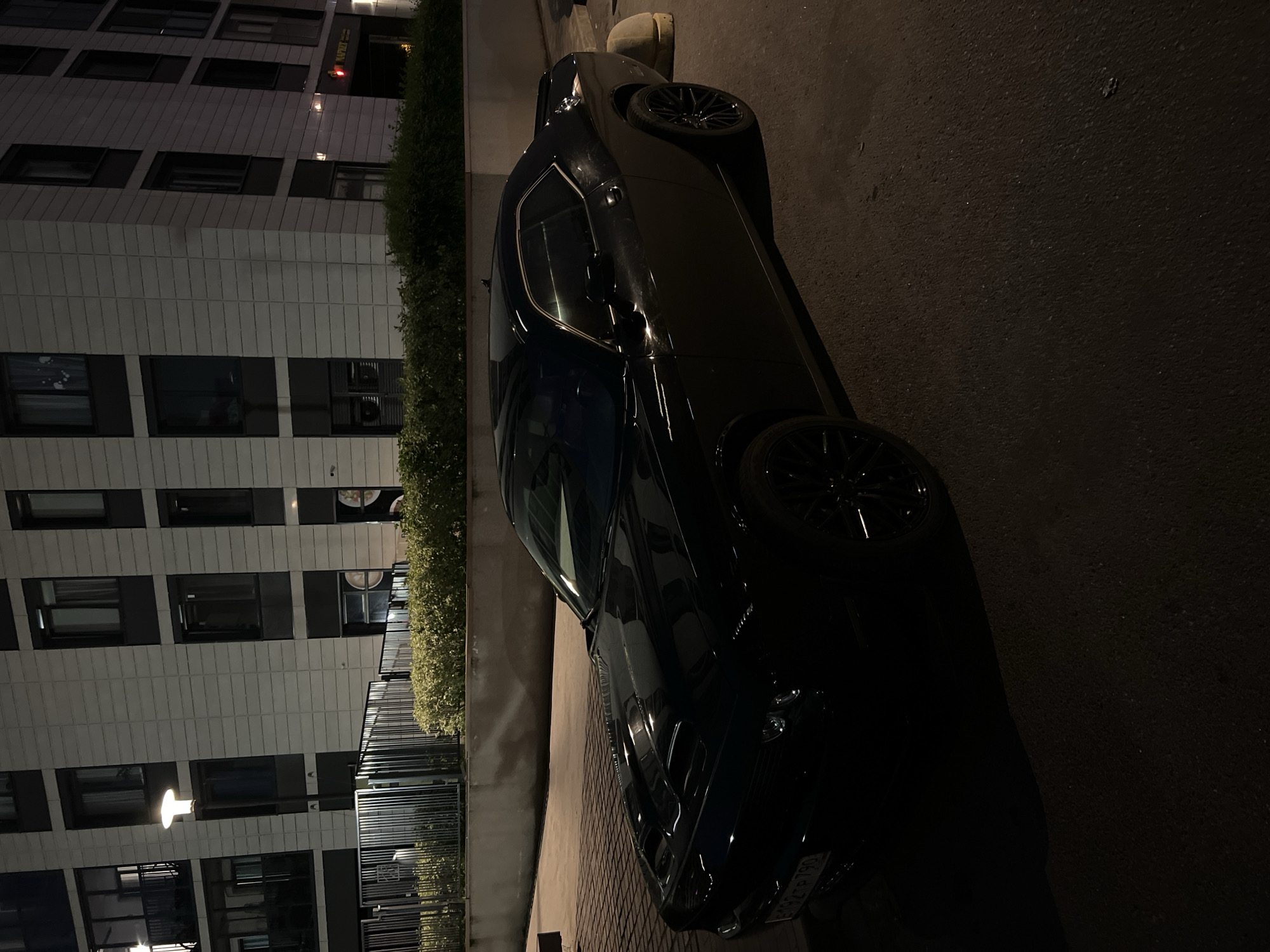 A black Dodge Challenger is parked on a paved surface in front of a multi-story building at night. The building has a modern facade with repetitive windows and dark accents. A row of manicured green bushes runs along the base of the building, separated from the parking area by a metal fence. The streetlights cast an ambient glow on the scene, highlighting the car's sleek design and the texture of the pavement. The license plate of the car is partially visible and appears to be a Russian plate. The overall scene depicts a quiet urban environment at night.