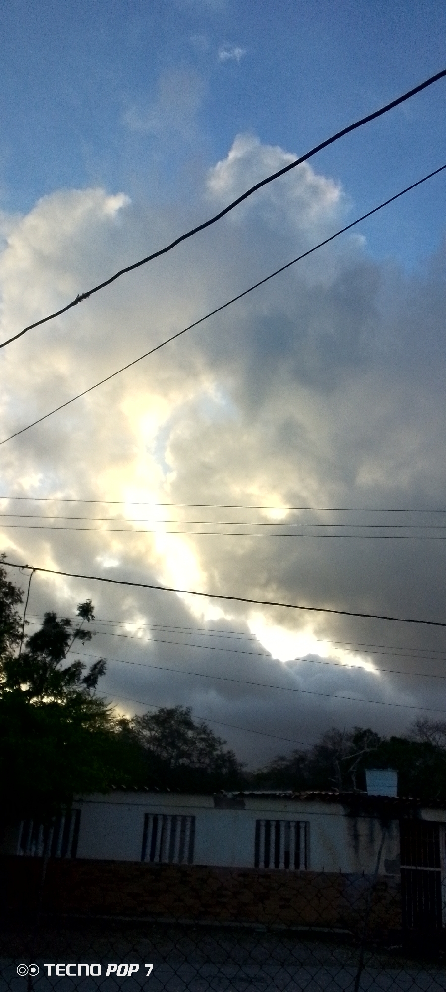 The image depicts a cloudy sky over a residential area, possibly in Altagracia, Venezuela. Several power lines traverse the frame horizontally. The sky is a mix of blue and dark gray clouds, with a bright, illuminated patch suggesting sunlight breaking through. In the foreground, a low-lying building with barred windows and a tiled roof is visible. Trees partially obscure the view of the building. A chain-link fence runs along the bottom of the image, and the text "TECNO POP 7" is visible in the lower left corner, likely a camera watermark. The scene appears to be captured during daylight, possibly late afternoon or early morning, given the lighting conditions.