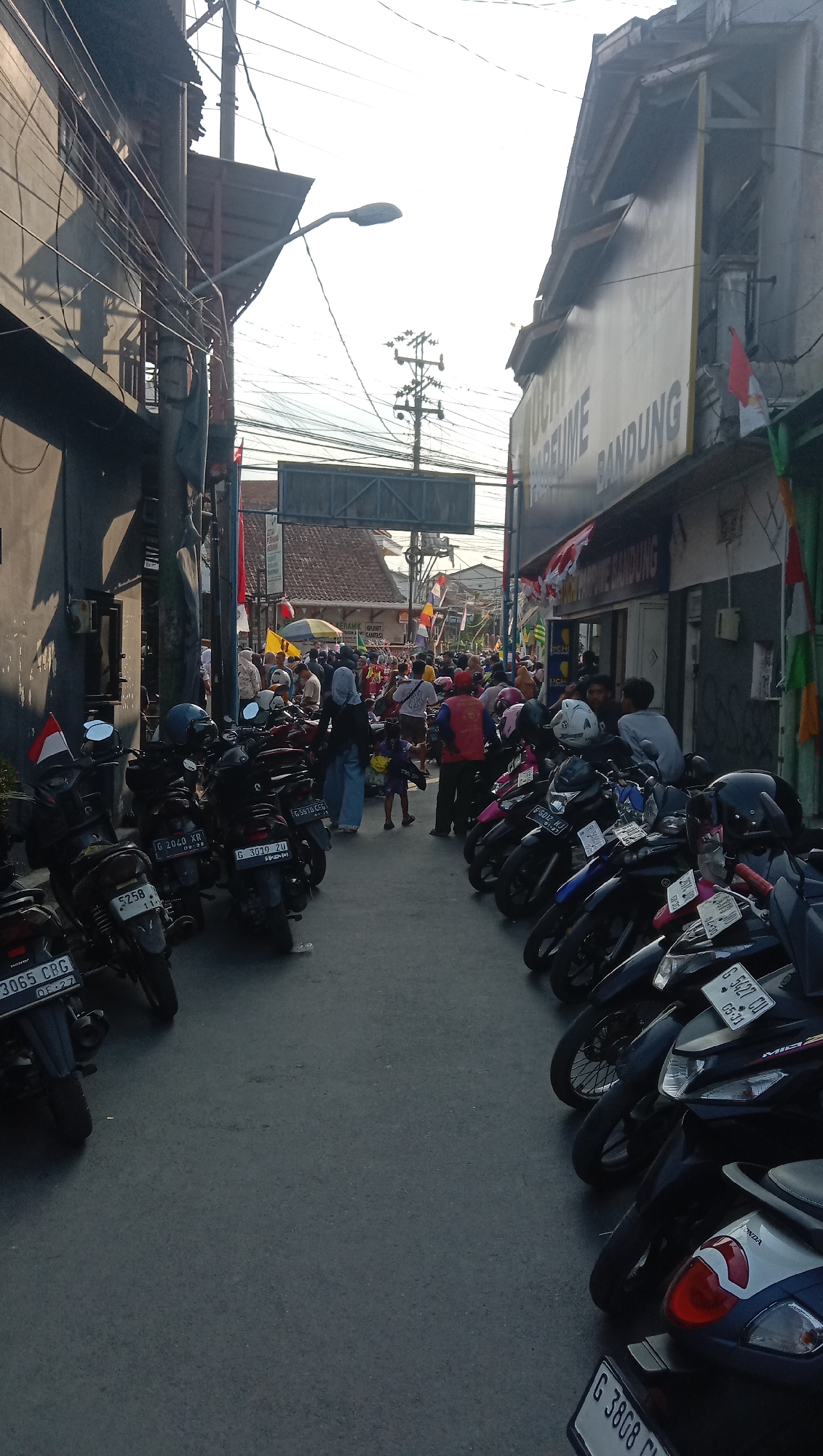 A street scene in Pesantren, Indonesia, is depicted under bright, hazy sunlight, casting long shadows. The street is lined with parked motorcycles on both sides, with a crowd of people in the middle, suggesting a busy market or event. Buildings flank the street, some with signs, including one reading "PARFUME BANDUNG." Red and white flags are visible, along with other colorful banners. The atmosphere appears to be that of a lively community gathering or commercial activity.