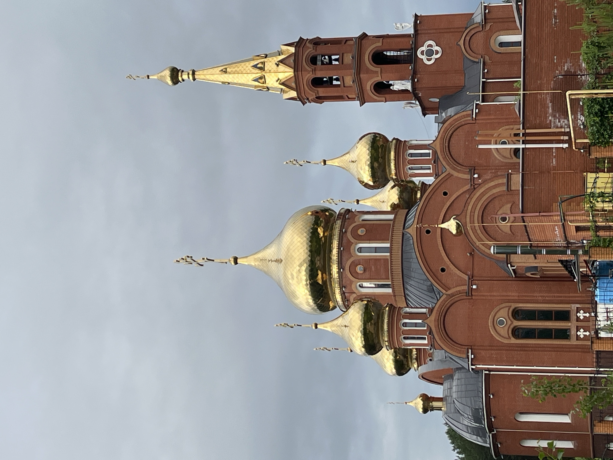 This is a vertical, eye-level, medium shot of the Church of the Holy Trinity in Chaikovsky, Russia. The red brick church features multiple golden domes topped with crosses. A tall, slender bell tower also adorned with gold is visible to the right of the main structure. The sky is overcast and gray, contrasting with the bright gold of the domes and tower. The church is surrounded by trees and vegetation, and a brick wall with fencing is in the foreground. No people are visible in the image.