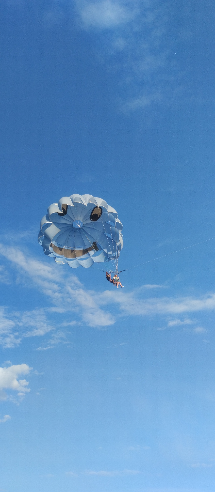 Two people are parasailing in a bright blue sky with scattered white clouds. The parasail is light blue and decorated with a smiling face. The people, one in a dark top and bottoms and the other in a lighter colored top and shorts, are suspended in the air below the parasail. The scene appears to be on a sunny day, typical of resort areas. The location is specified as Cholpon-Ata, Kyrgyzstan, a known tourist destination on the shores of Lake Issyk-Kul.