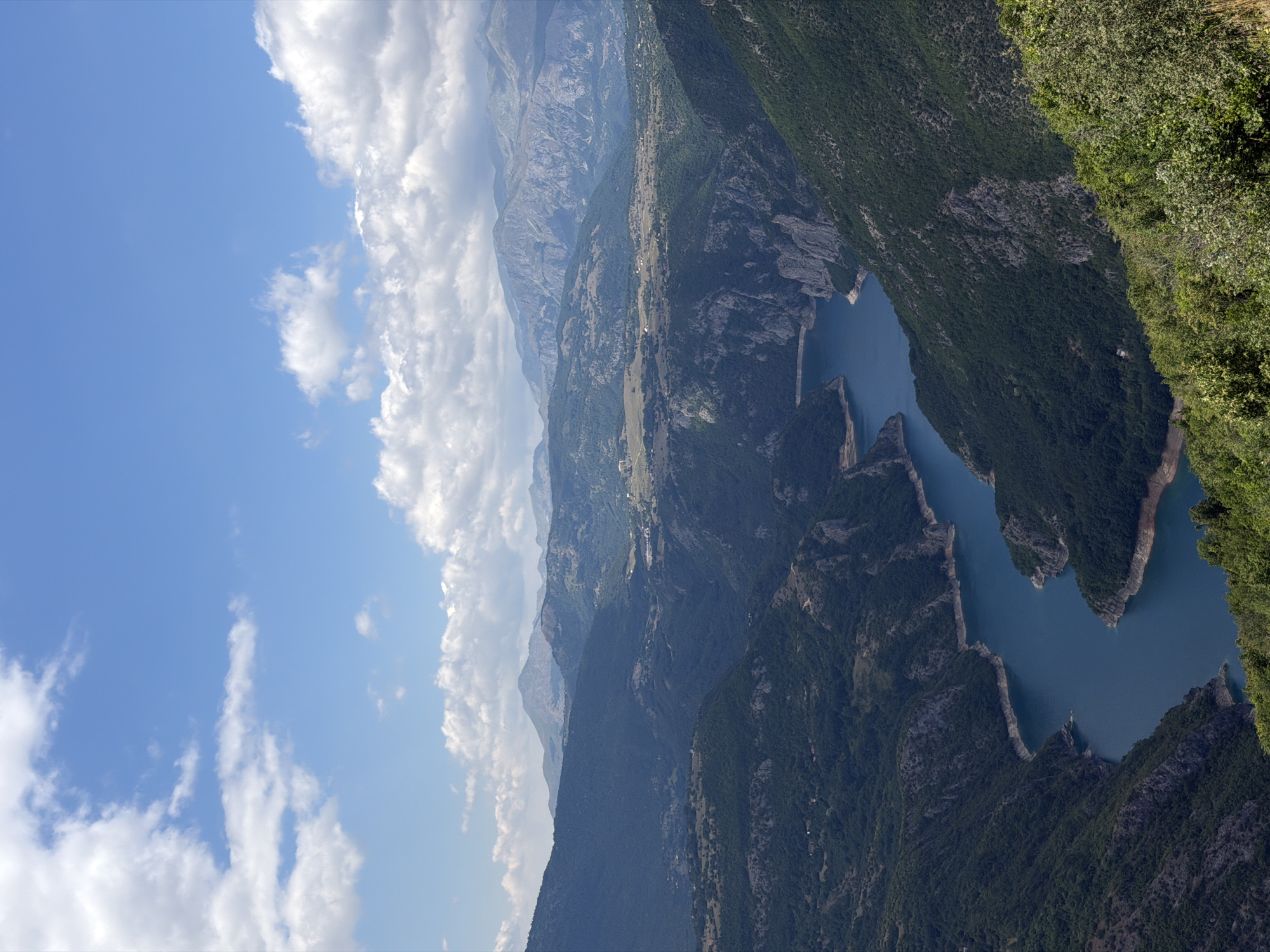 The image shows a panoramic view of a deep canyon with a winding, blue river flowing through it. The canyon walls are steep and covered with lush green trees. In the distance, several layers of mountains are visible, with the furthest ranges appearing hazy. The sky is blue with scattered white clouds. This scene is located near the city of Dubljevići, Montenegro, specifically depicting the Piva River Canyon. The image does not contain any people, objects, or actions.