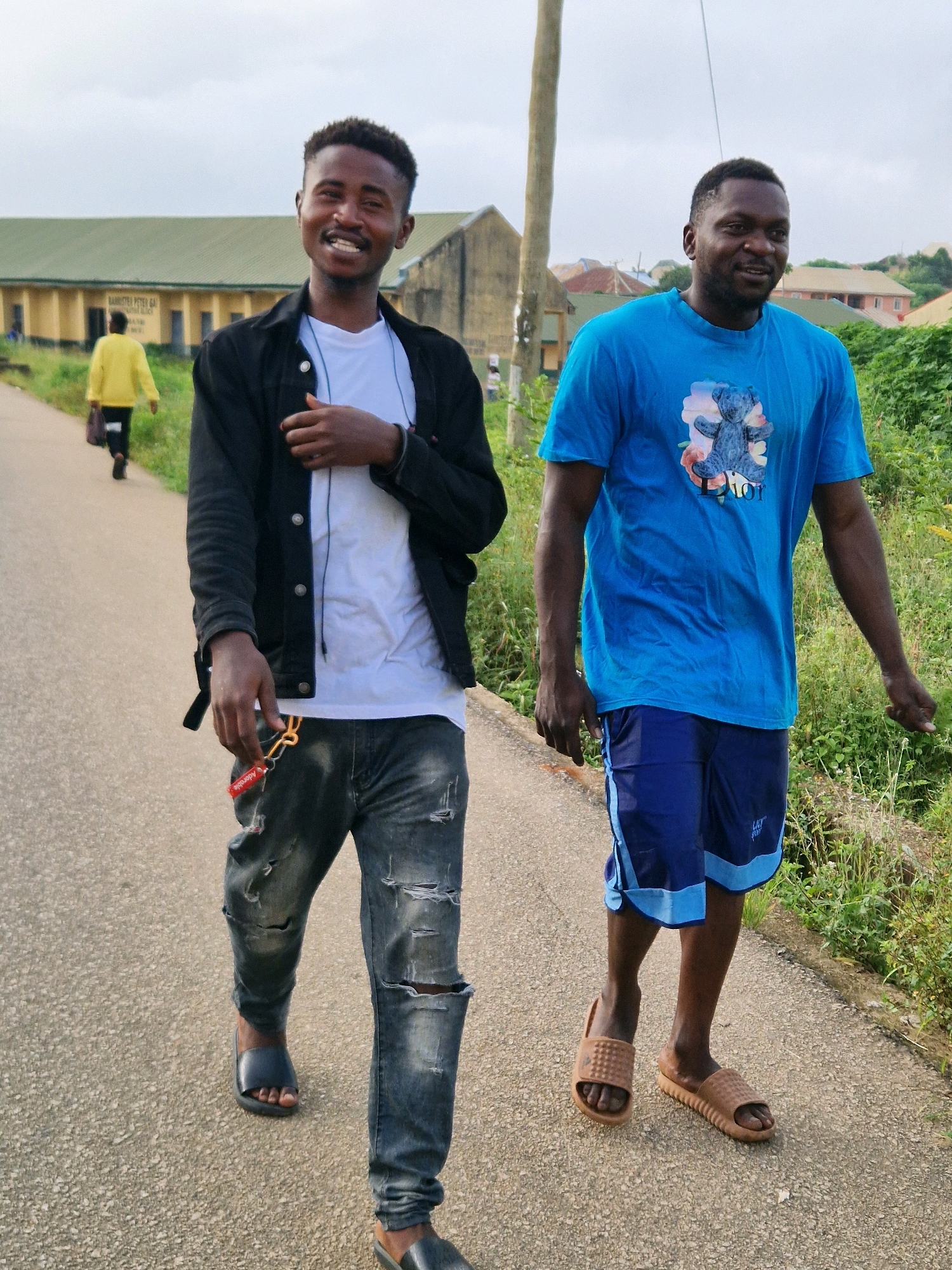 The image shows two young men walking on a paved road in Kabong, Nigeria. The man on the left is wearing a black jacket over a white t-shirt and ripped denim jeans, with black sandals. He is smiling and looking at the camera, with a keychain with a red tag in his hand. The man on the right is wearing a blue t-shirt with a graphic design and blue athletic shorts with light blue stripes, with brown slides. He is looking to his right and smiling. In the background, a person in a yellow jacket is walking away from the camera on the same road. Further in the background, there are buildings with light green roofs and some vegetation. The sky is overcast.