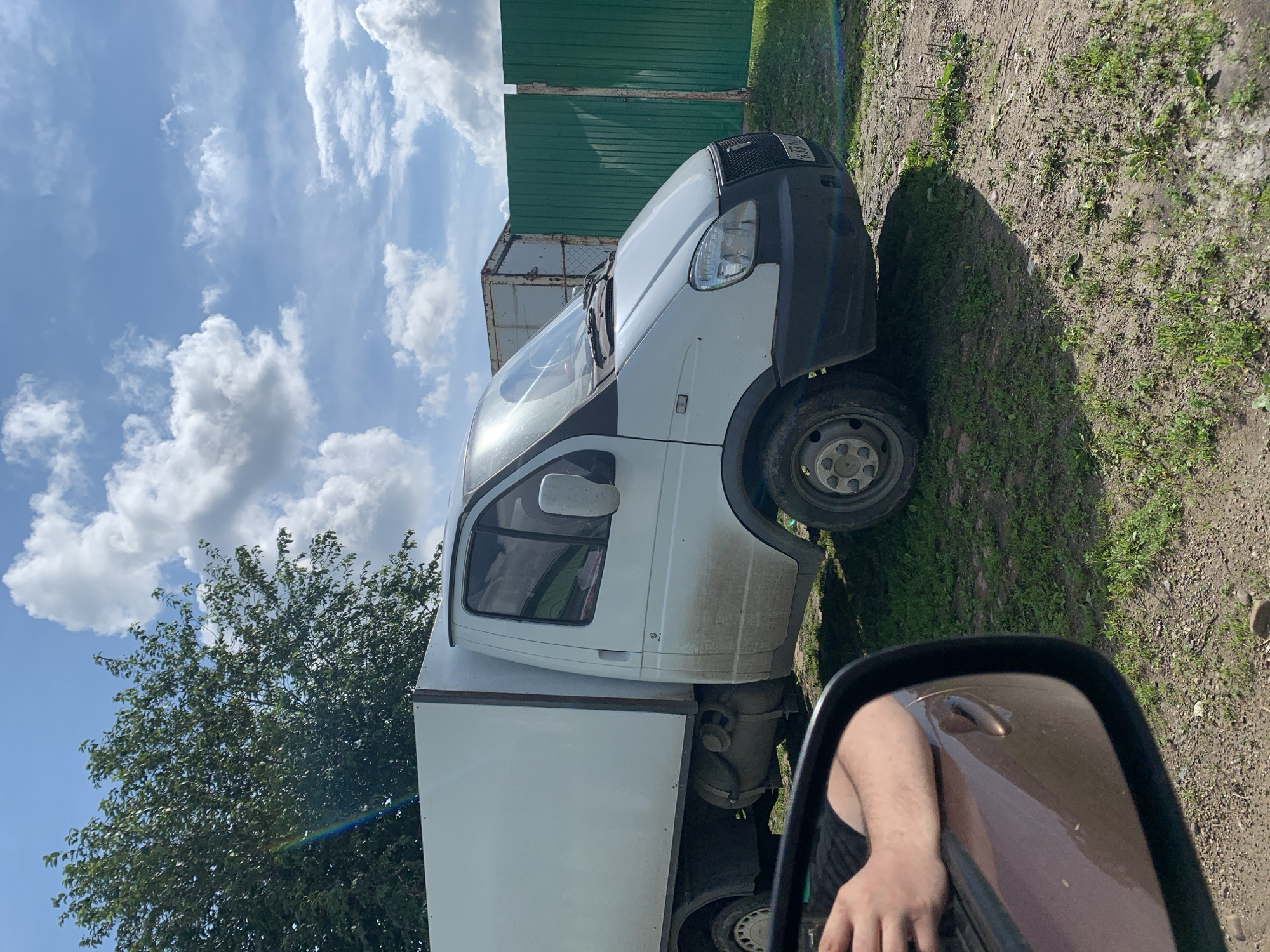 In Barsai, Russia, on a sunny day with scattered white clouds, a white delivery van with a cargo box is parked on a dirt and grass lot. The van is dirty, with mud splattered on its lower panels and wheels. A green corrugated metal fence stands to the right of the van, and a rusted shipping container is visible behind it. In the foreground, a side-view mirror of a car shows a person's arm and shoulder, indicating the photo was taken from inside another vehicle. A large tree with green leaves is on the left side of the frame, and a subtle rainbow arc is visible near the top left corner. The ground is a mix of gravel, dirt, and sparse green grass.