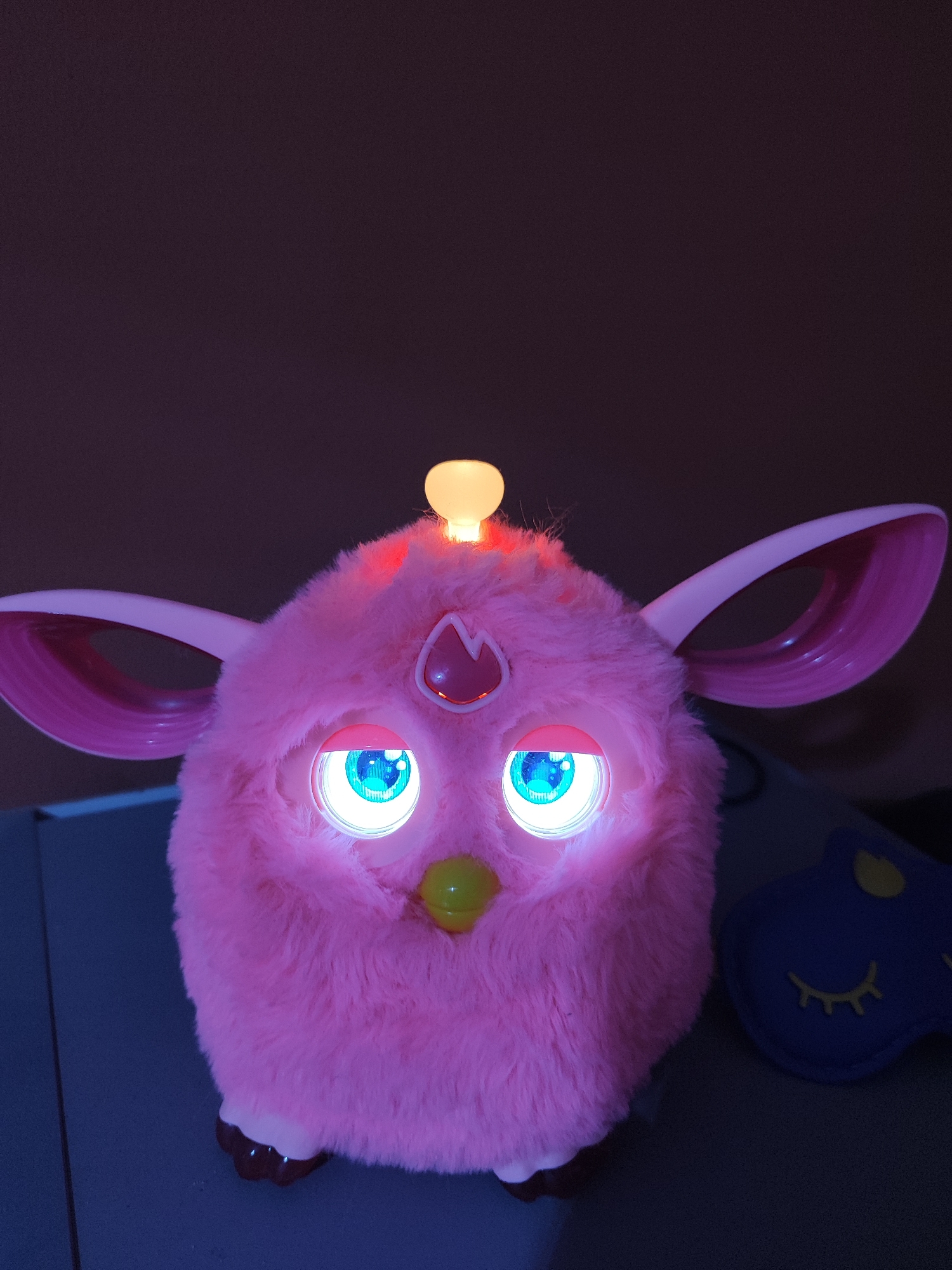 A pink, fluffy toy with large, luminous blue eyes sits on a dark surface. The toy has large, pink, oval-shaped ears and a small, yellow beak. A light bulb-shaped object is illuminated on top of its head, and a red gem-like shape is visible on its forehead. A blue sleep mask with yellow sleepy eyes is placed to the right of the toy. The background is a dark, blurred purple. The image is taken indoors, likely in a room with low lighting. There is no indication of the city of May, Uzbekistan within the image.