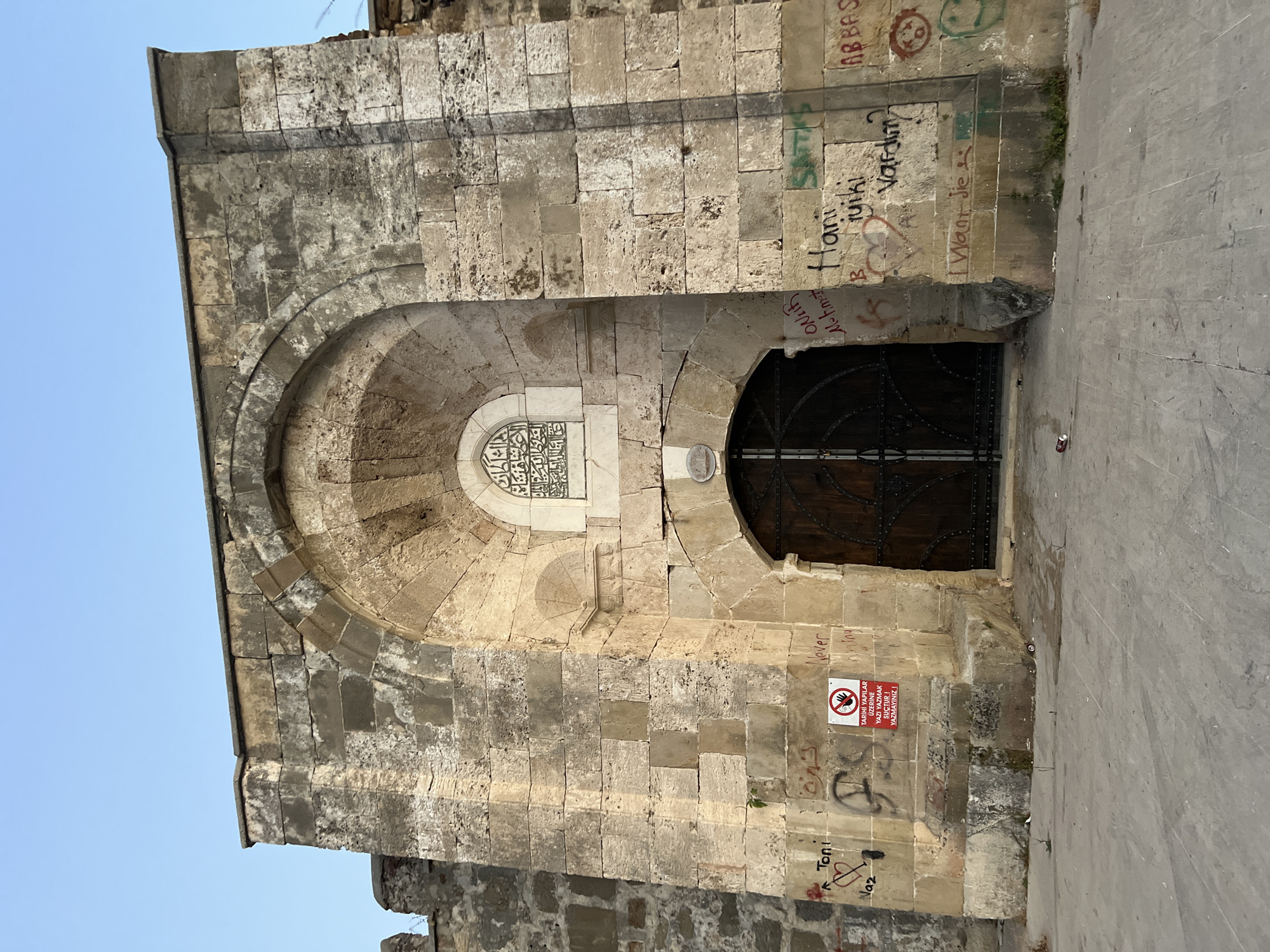 The image shows the exterior of an ancient stone structure, likely a gate or entrance, located in Alanya, Turkey. The architecture features rough-hewn stone blocks and a prominent arched opening. Above the arched entrance, there is a smaller, rectangular section with Arabic calligraphy. The stone surfaces are weathered and show signs of age.
The structure is covered in graffiti, with various names and messages scrawled in different colors and styles. Some of the graffiti includes names like "Toni," "Naz," "Mohmet," "Hani," "Ruiki," and "ABBAS." There is also a red and white sign with a hand symbol and Turkish text, which reads "TARİHİ YAPILAR ÜZERİNE YAZMAK YASAKTIR! SUÇTUR!" (It is forbidden to write on historical buildings! It is a crime!). A red heart symbol is also visible.
The ground in front of the structure is paved with large stone slabs, and there is some sparse vegetation growing near the base of the wall. The sky above is clear and blue. The lighting suggests it is daytime, with the sun casting shadows. There are no people visible in the image. The overall impression is one of historical significance marred by modern vandalism.