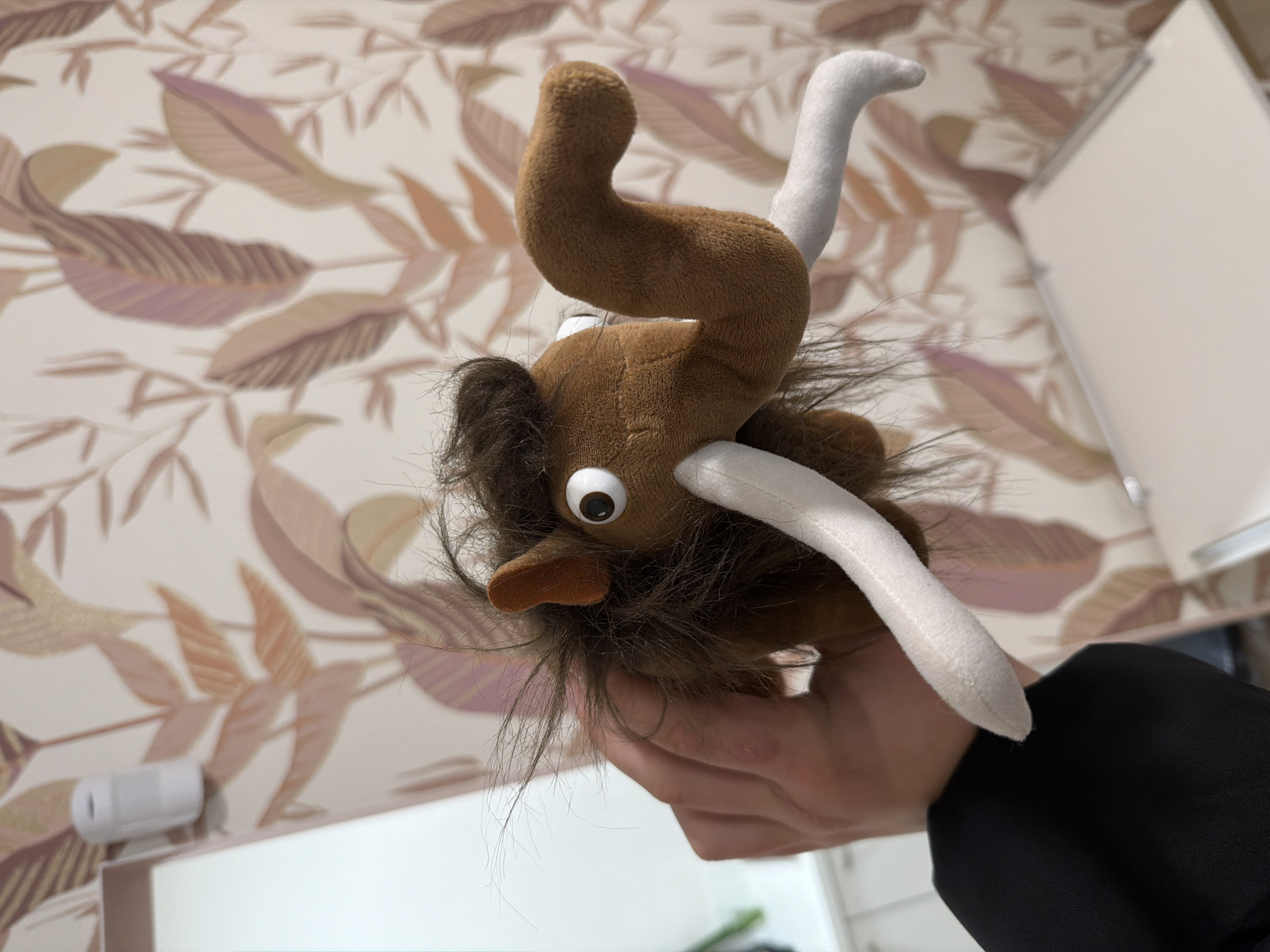 A brown plush mammoth toy is being held up in front of a wall decorated with a leaf pattern. The toy has white tusks and shaggy brown fur around its face. A hand wearing a black sleeve is holding the toy. The background includes a white object (possibly a light switch or sensor) on the wall and a white cabinet or door to the right. There is no information in the image to determine the location in Burgas, Bulgaria.