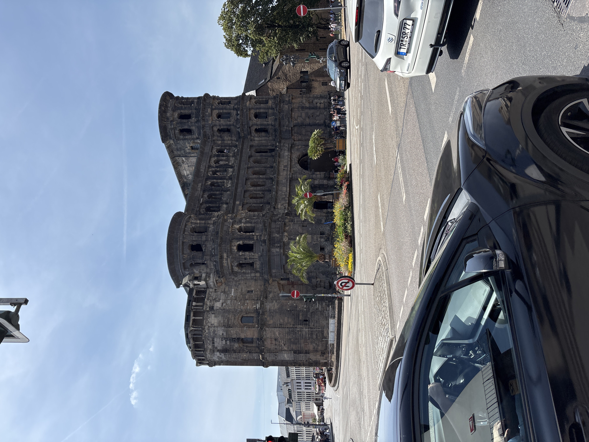 The image shows the Porta Nigra in Trier, Germany, on a clear, sunny day. The ancient Roman gate, constructed from dark stone blocks, dominates the center of the frame. Its multi-tiered structure features arched openings and ornate stonework. In front of the gate, a circular garden with colorful flowers and palm trees is visible.
A street intersection is in the foreground with several cars. A black car is closest to the camera on the left, with its driver's arm visible. To the right, a white Suzuki is parked in a lane. A dark gray SUV is turning in the intersection. Traffic lights and road signs are present, including a "no U-turn" sign.
Pedestrians are walking near the Porta Nigra and on the sidewalk in the background. Buildings with traditional European architecture line the street to the left of the gate. The sky is blue with a few wispy clouds. The overall scene depicts a vibrant urban environment with a significant historical landmark.