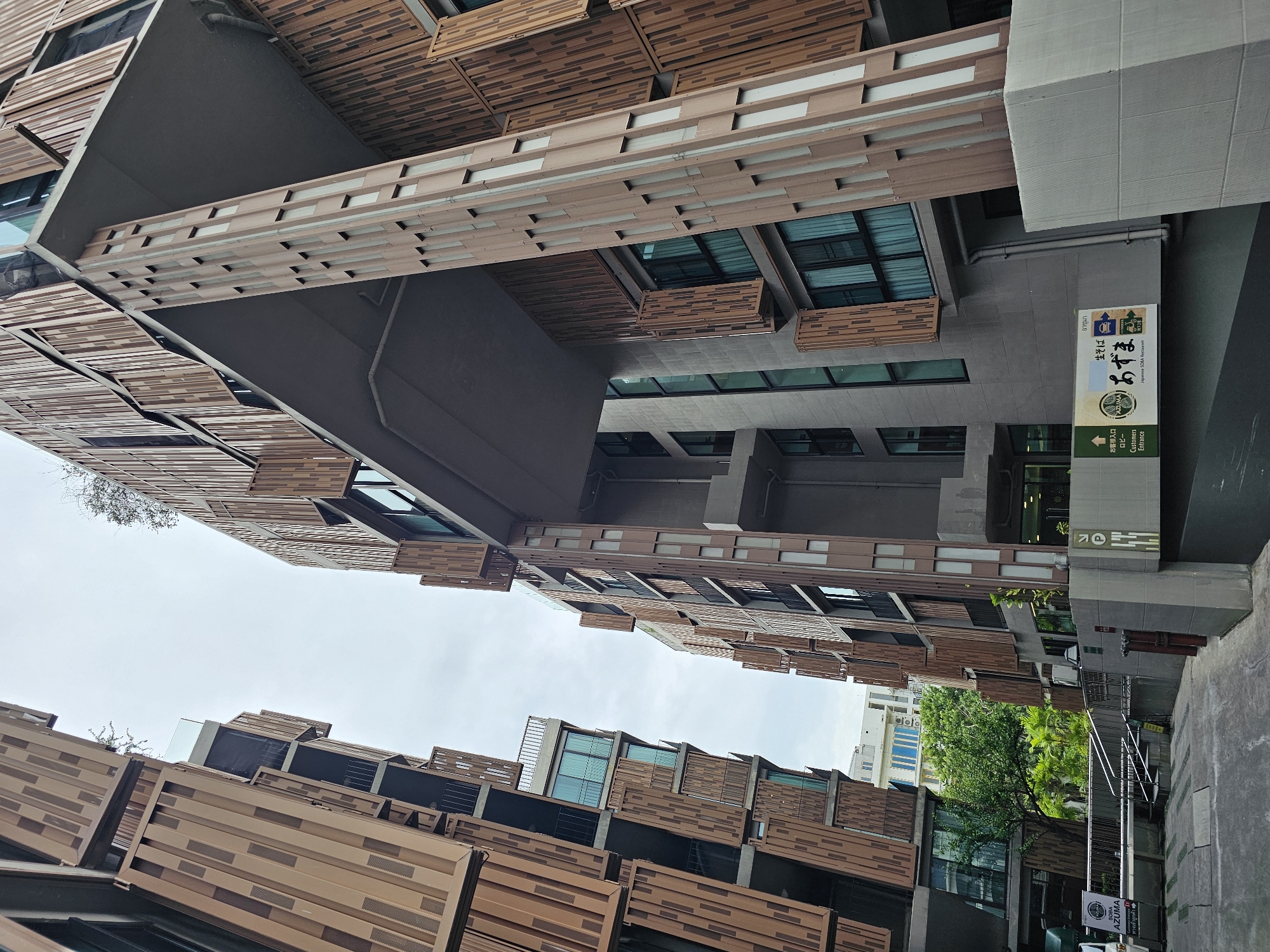 The image shows the exterior of a modern apartment building in Bangkok, Thailand. The building features a unique design with alternating wooden louvers and windows on its facade. Two sections of the building are visible, angled towards each other, creating a sense of depth. One section is closer to the foreground and appears to be a corner of the building, with a large, angled concrete support structure. The other section is further back and extends upwards. The sky is overcast. In the foreground, there is a paved walkway and some greenery. A sign with Japanese text and an arrow pointing to "Customers Entrance" is visible on the left. Another sign with Japanese characters, including "SOBA" and "AZUMA," and a car icon, indicating a "Japanese SOBA Restaurant," is also present. The overall impression is of a stylish, contemporary urban dwelling.