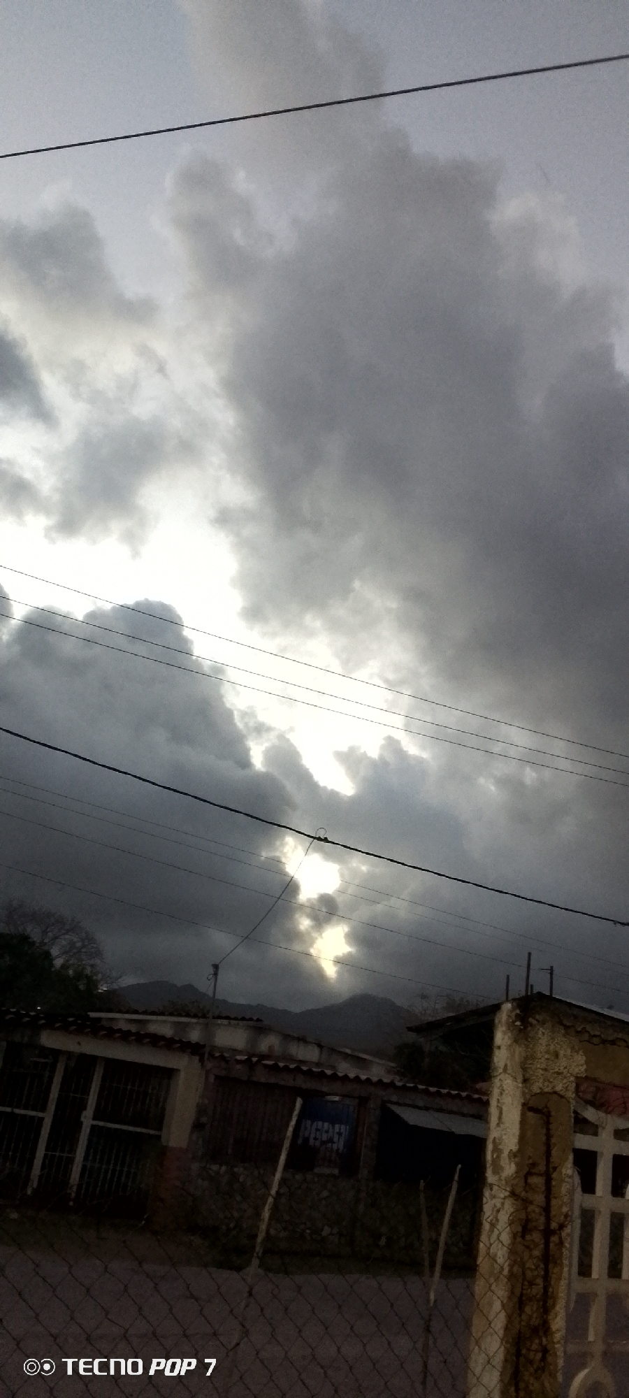 The image presents a cloudy sky above a street scene in Altagracia, Venezuela. The sky is heavily overcast with dark gray clouds, with a sliver of light breaking through. Power lines stretch across the sky. Below, a low-slung building with a corrugated metal roof is visible. The building has a Pepsi advertisement on its side. A chain-link fence and a weathered concrete pillar stand in the foreground. In the distance, a faint mountain range can be seen. The image appears to be taken in the early morning or late evening, given the dim lighting conditions. A TECNO POP 7 watermark is visible in the bottom left corner.